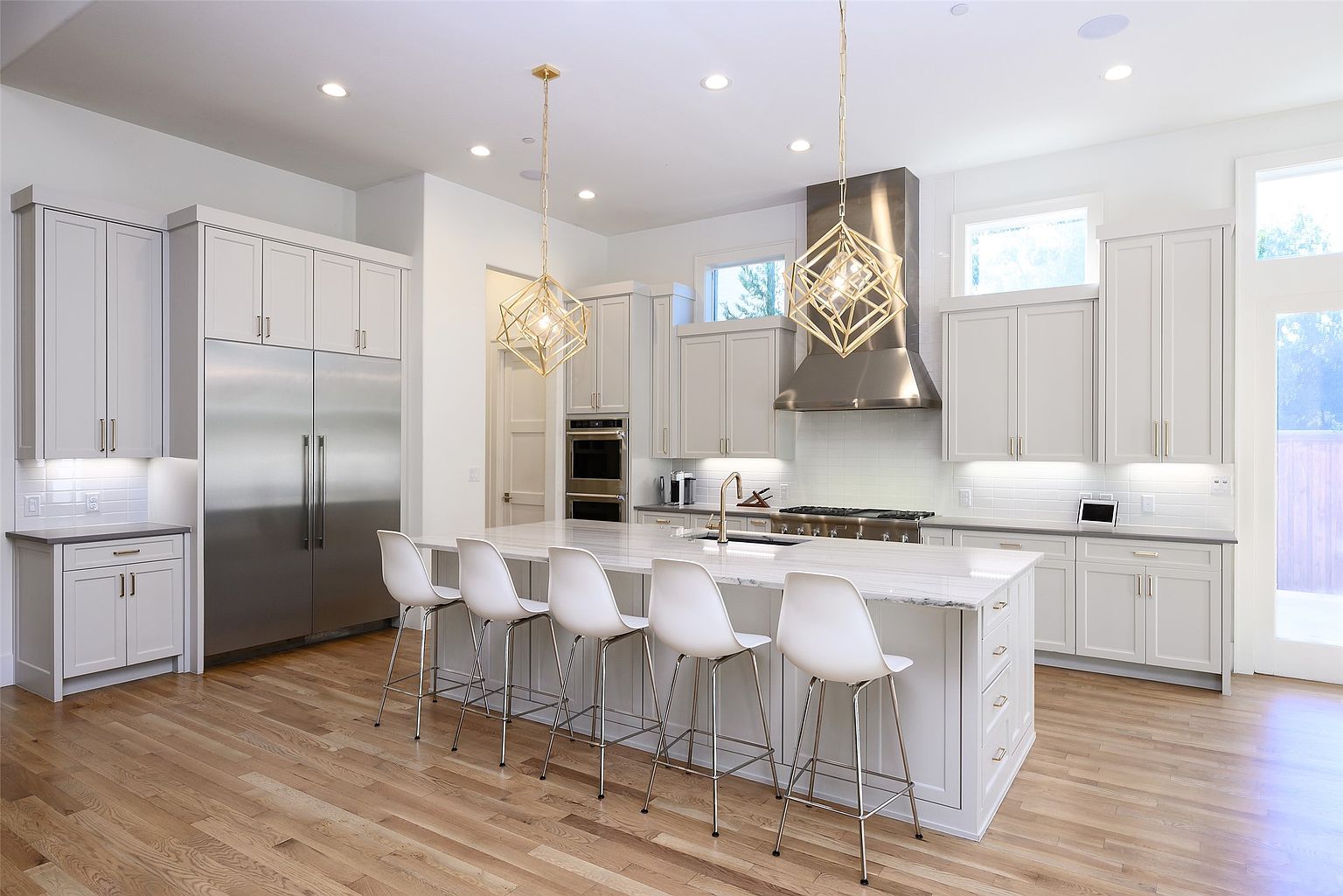This is a bright and spacious kitchen featuring white cabinetry, stainless steel appliances, and a large center island with a marble countertop. The kitchen is illuminated by pendant lighting and recessed lights, creating a warm and inviting atmosphere. The hardwood floors add a touch of elegance to the space, and the overall design is modern and sophisticated.