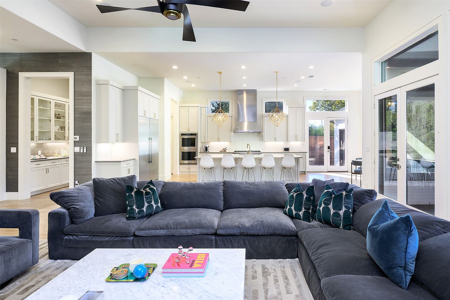 This is a spacious and modern living room that seamlessly flows into the kitchen area. The living room features a large, comfortable gray sectional sofa with decorative pillows, a marble coffee table, and a neutral-toned rug. The open floor plan allows for easy interaction between the living and kitchen spaces, creating an inviting atmosphere.