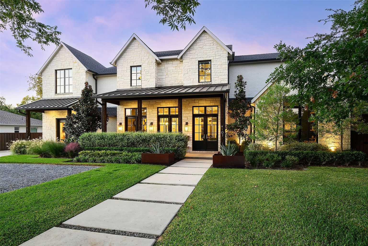 This is a front exterior view of a large, luxurious two-story home. The house features a combination of stone and painted brick facade, with black-framed windows and a covered porch supported by dark wood columns. A well-manicured lawn and a stone pathway lead to the front entrance, enhancing the property's curb appeal.