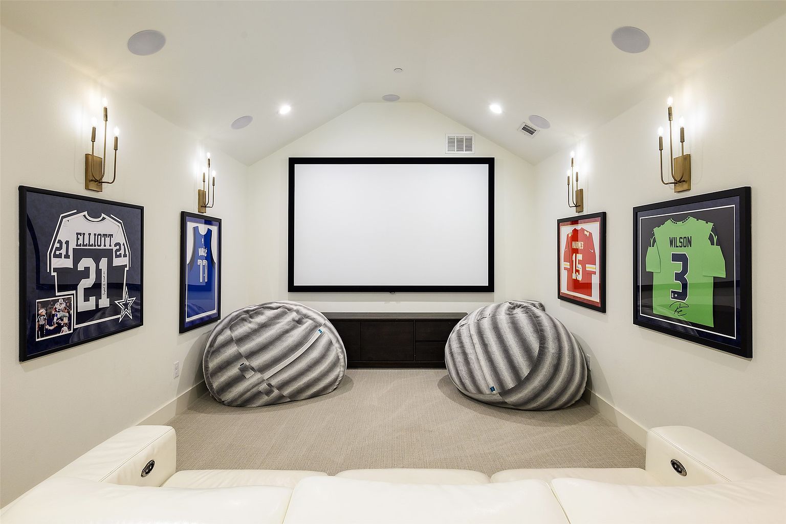 This is an interior shot of a home theater room. The room features a large projection screen, two beanbag chairs, and a white leather sofa. Sports memorabilia adorns the walls, and sconces provide ambient lighting, creating a cozy and inviting atmosphere.