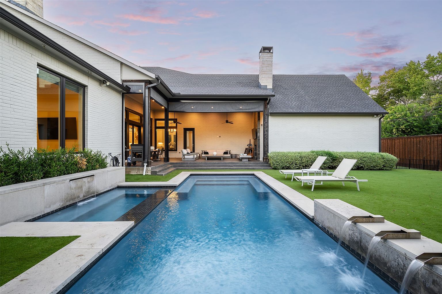 This image showcases a luxurious backyard pool area. The rectangular pool features modern design elements, including integrated water features and sleek coping. Two lounge chairs are placed on the well-maintained lawn, and an open-air patio area with comfortable seating is visible in the background, creating an inviting outdoor living space.