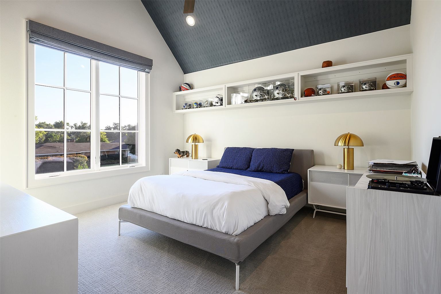 This is a well-lit bedroom featuring a gray platform bed with white bedding and blue pillows. The room has a modern aesthetic with white walls, a dark blue ceiling, and built-in shelving displaying sports memorabilia. Natural light floods the space through a large window, creating a bright and inviting atmosphere.