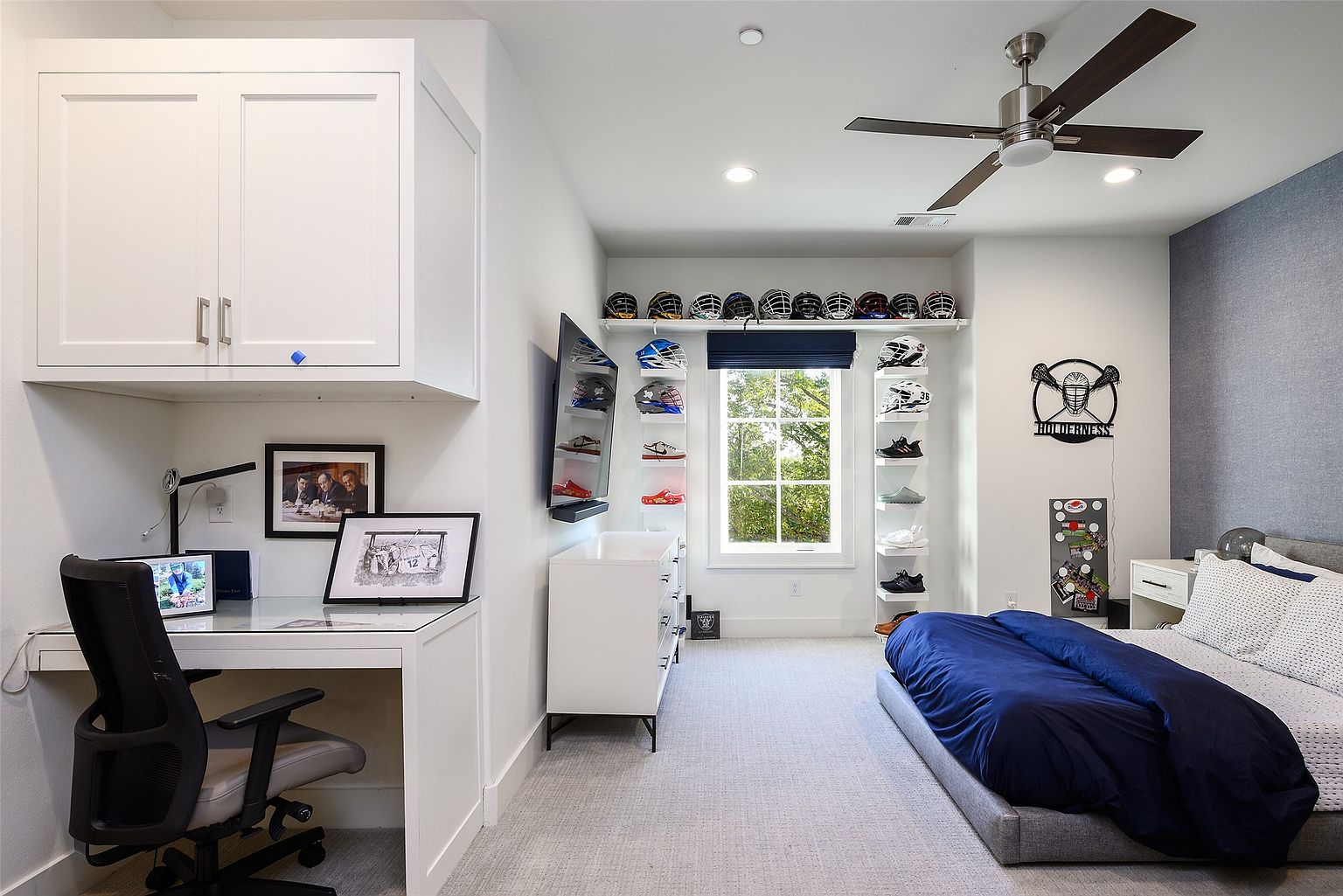 This is a well-organized bedroom featuring a built-in desk area with white cabinetry, a bed with a navy blue comforter, and shelving displaying sports helmets and shoes. The room has a modern aesthetic with a neutral color palette, complemented by a ceiling fan and a window providing natural light. The room is designed with a focus on storage and display, creating a functional and stylish space.