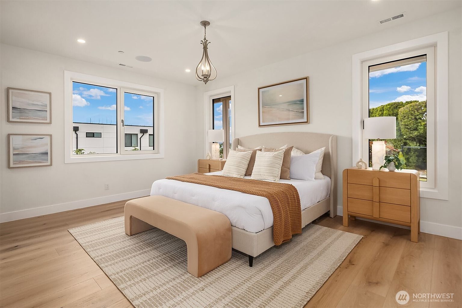 This bright and airy primary bedroom features light wood flooring, neutral walls, and a large upholstered bed centered in the room. The space is accented by a textured area rug, a matching bench at the foot of the bed, and two light wood nightstands topped with modern lamps. Large windows on multiple walls provide ample natural light and views of the surrounding landscape, creating a serene and inviting atmosphere.