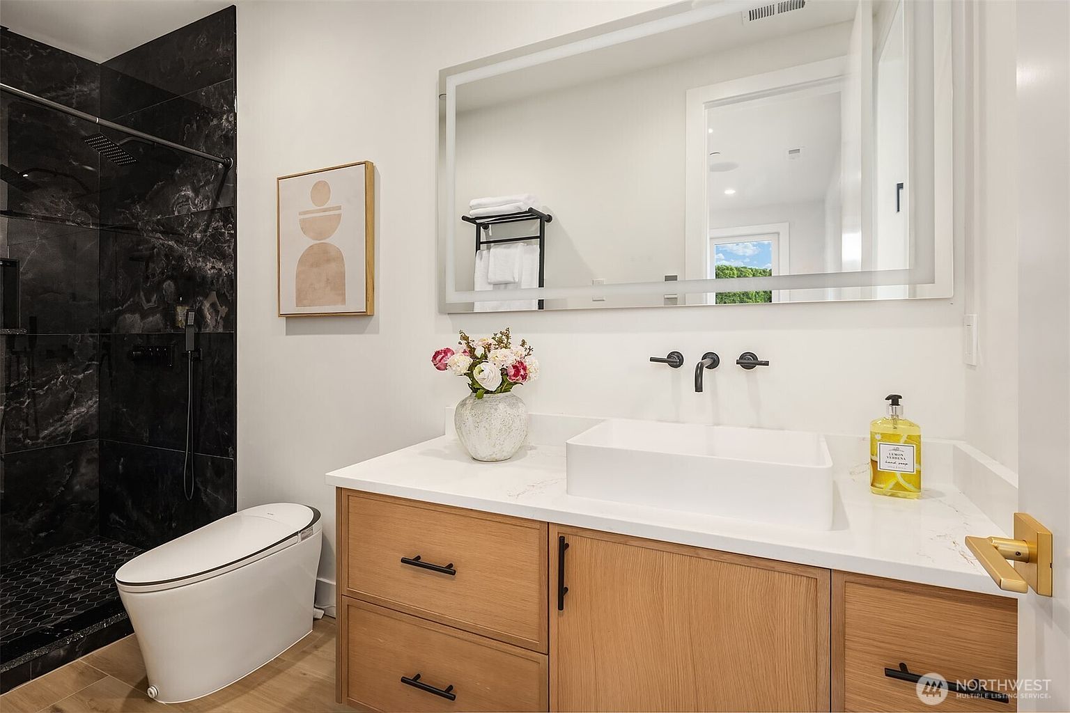 This modern bathroom features a sleek vanity with light wood cabinetry, a white rectangular vessel sink, and a wall-mounted faucet. To the left, a walk-in shower is enclosed by black-framed glass and finished with dark, textured stone tiles. A large, rectangular mirror with integrated lighting hangs above the vanity, reflecting the bright, clean aesthetic of the space.