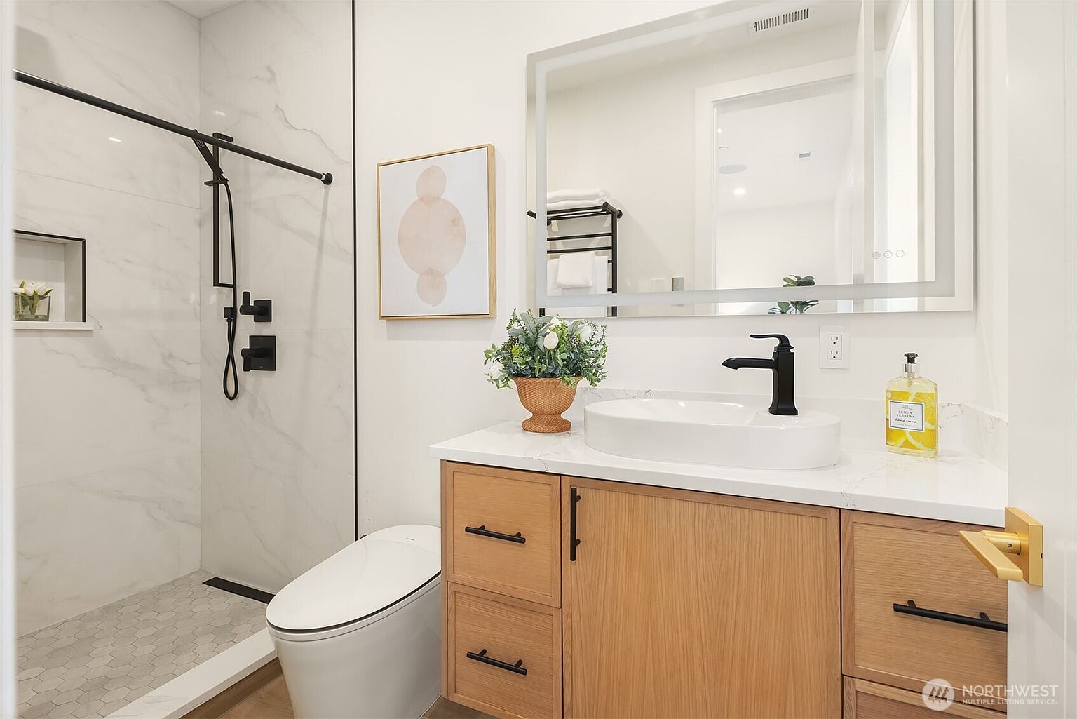 This modern bathroom features a light wood vanity with a white countertop and a vessel sink, complemented by matte black fixtures. To the left, a walk-in shower is enclosed by a glass panel and finished with light-colored, veined wall tiles and a hexagonal floor tile pattern. A large, rectangular LED-lit mirror hangs above the vanity, reflecting the clean, contemporary aesthetic of the space.