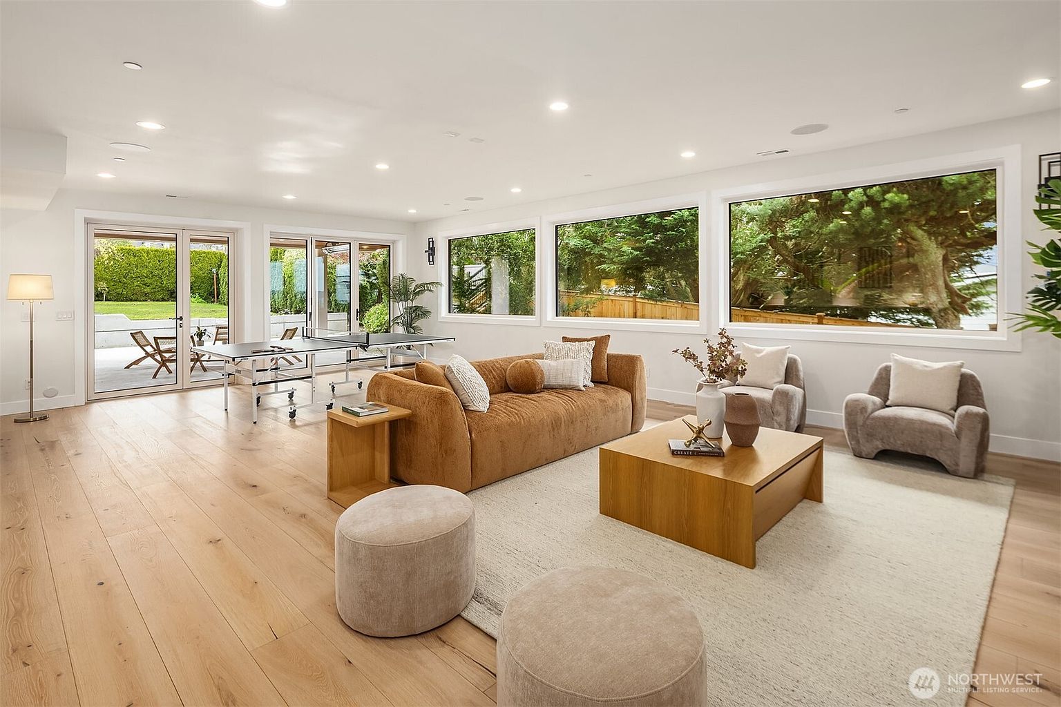 This spacious, light-filled living room features light wood flooring and a neutral color palette, anchored by a large, textured brown sofa and a matching wooden coffee table. The room seamlessly transitions into a recreational area with a ping-pong table near the glass doors that open to an outdoor patio. Large windows along the back wall provide expansive views of the lush greenery outside, creating a bright and airy atmosphere perfect for relaxation or entertainment.