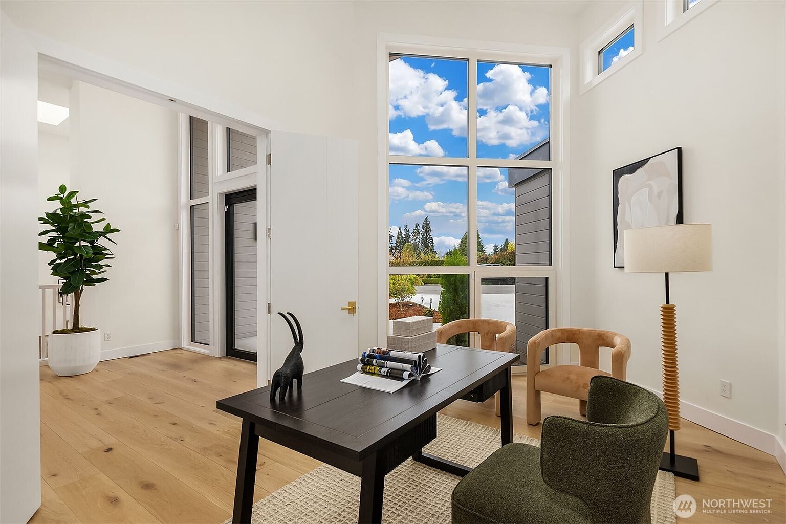 This bright and modern home office features light wood flooring and a large, dark-toned desk positioned to face a wall of tall windows that provide ample natural light. The room is furnished with two tan armchairs and a green accent chair, creating a comfortable seating area, while a tall floor lamp and a piece of abstract wall art add a touch of sophistication. The space feels open and airy, with a view into an adjacent hallway and a potted plant adding a touch of greenery to the minimalist aesthetic.