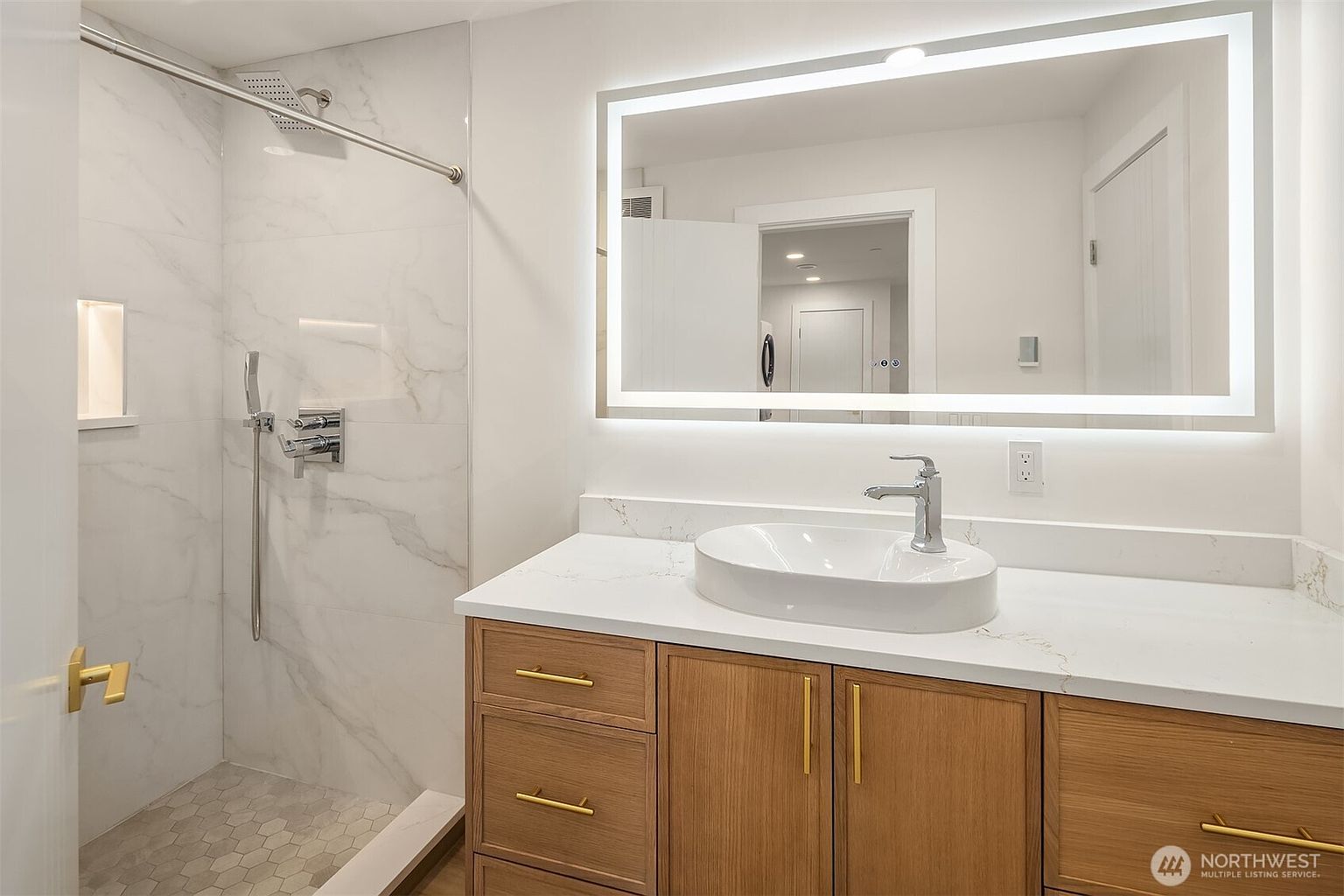 This modern bathroom features a light wood vanity with gold-toned hardware and a white countertop topped with a vessel sink. To the left, a walk-in shower is enclosed by a glass panel and finished with light-colored stone wall tiles and a hexagonal tile floor. A large, rectangular LED-backlit mirror hangs above the vanity, creating a bright and contemporary aesthetic.