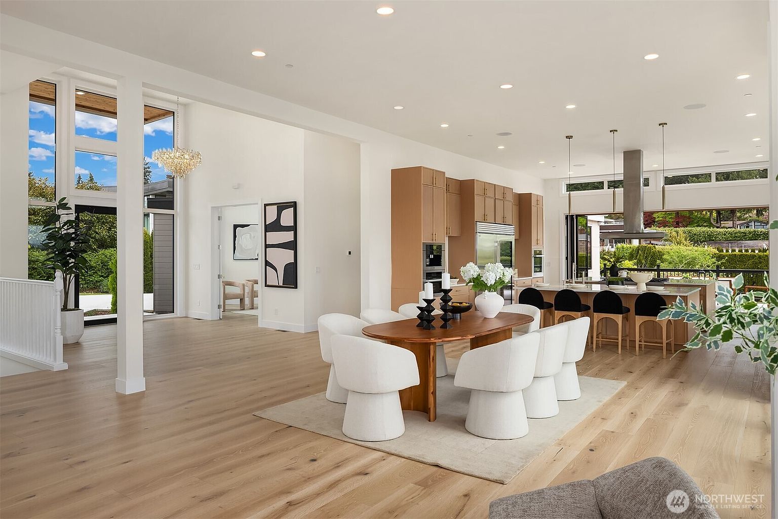 This open-concept living space features a dining area with a wooden table surrounded by six modern, curved white chairs on a neutral area rug. Beyond the dining area, a sleek kitchen with light wood cabinetry and a large island with bar seating is visible, leading to an outdoor patio through wide glass doors. The room is bright and airy, characterized by light wood flooring, recessed lighting, and a tall, modern chandelier near the entryway.