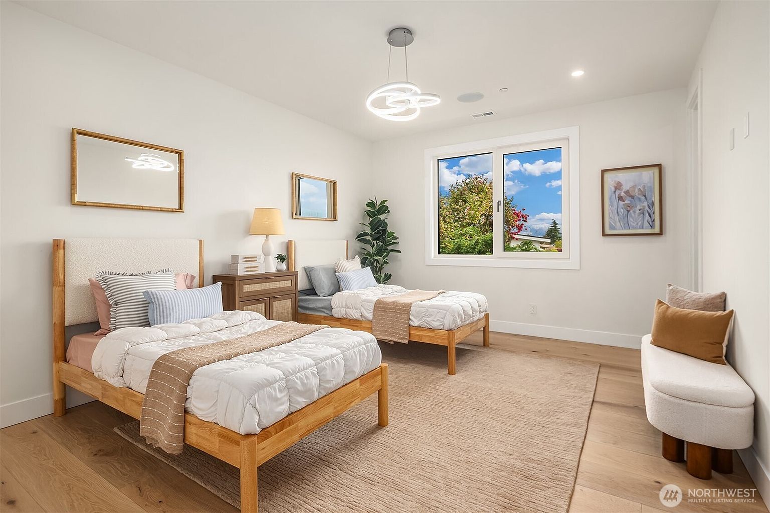 This bright and airy bedroom features two twin beds with light wood frames, dressed in white bedding with neutral-toned accents. The room is decorated with a minimalist aesthetic, including a central light fixture, a small nightstand, and a curved bench at the foot of the room. A large window allows for ample natural light, highlighting the light wood flooring and clean white walls.