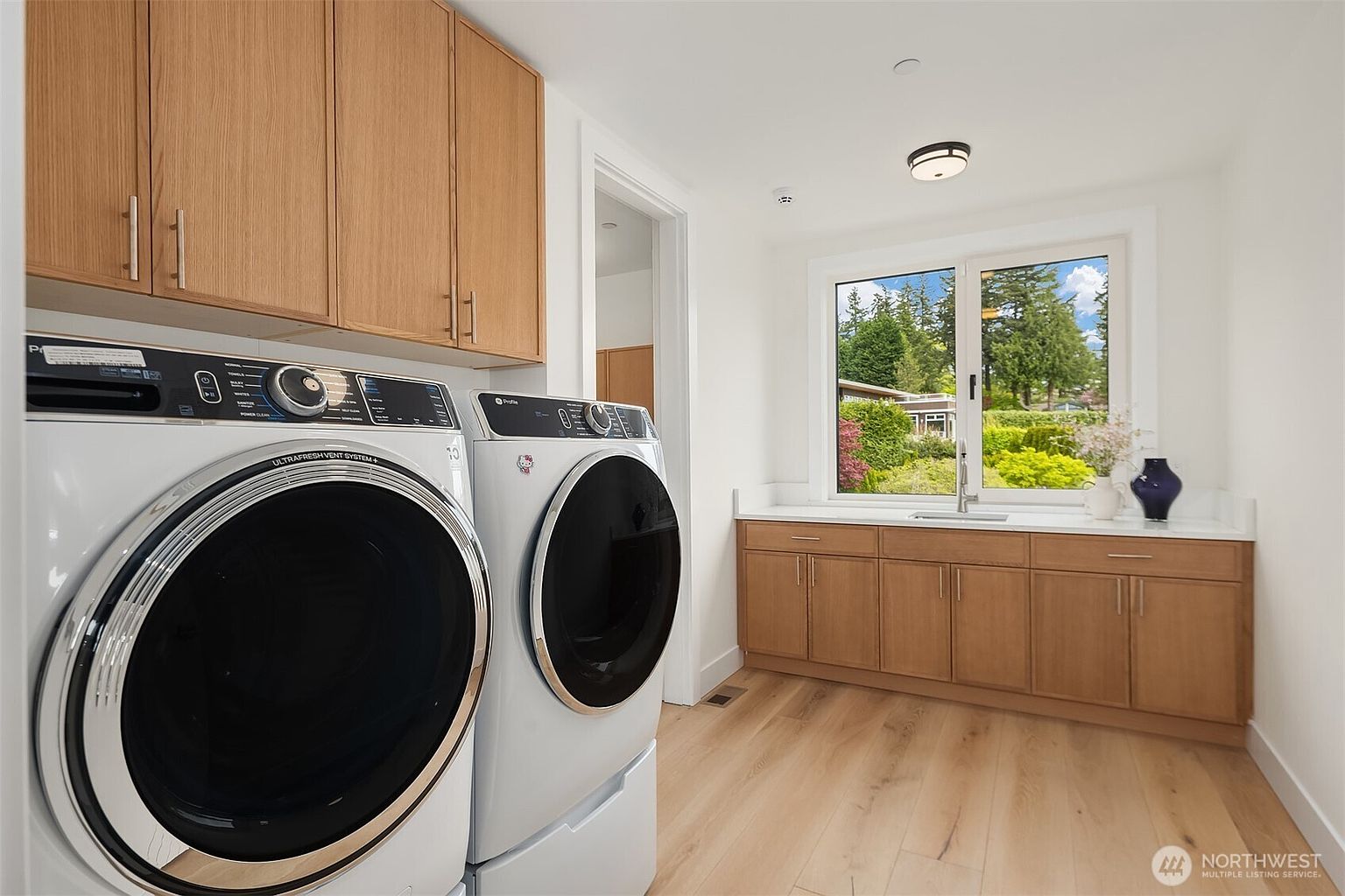 This bright and functional laundry room features light wood cabinetry providing ample storage above and below the counter space. A pair of modern white front-loading appliances sits prominently on the left, while a sink is integrated into the counter beneath a window that offers a view of the outdoors. The room is finished with light-toned wood flooring, creating a clean and cohesive aesthetic.