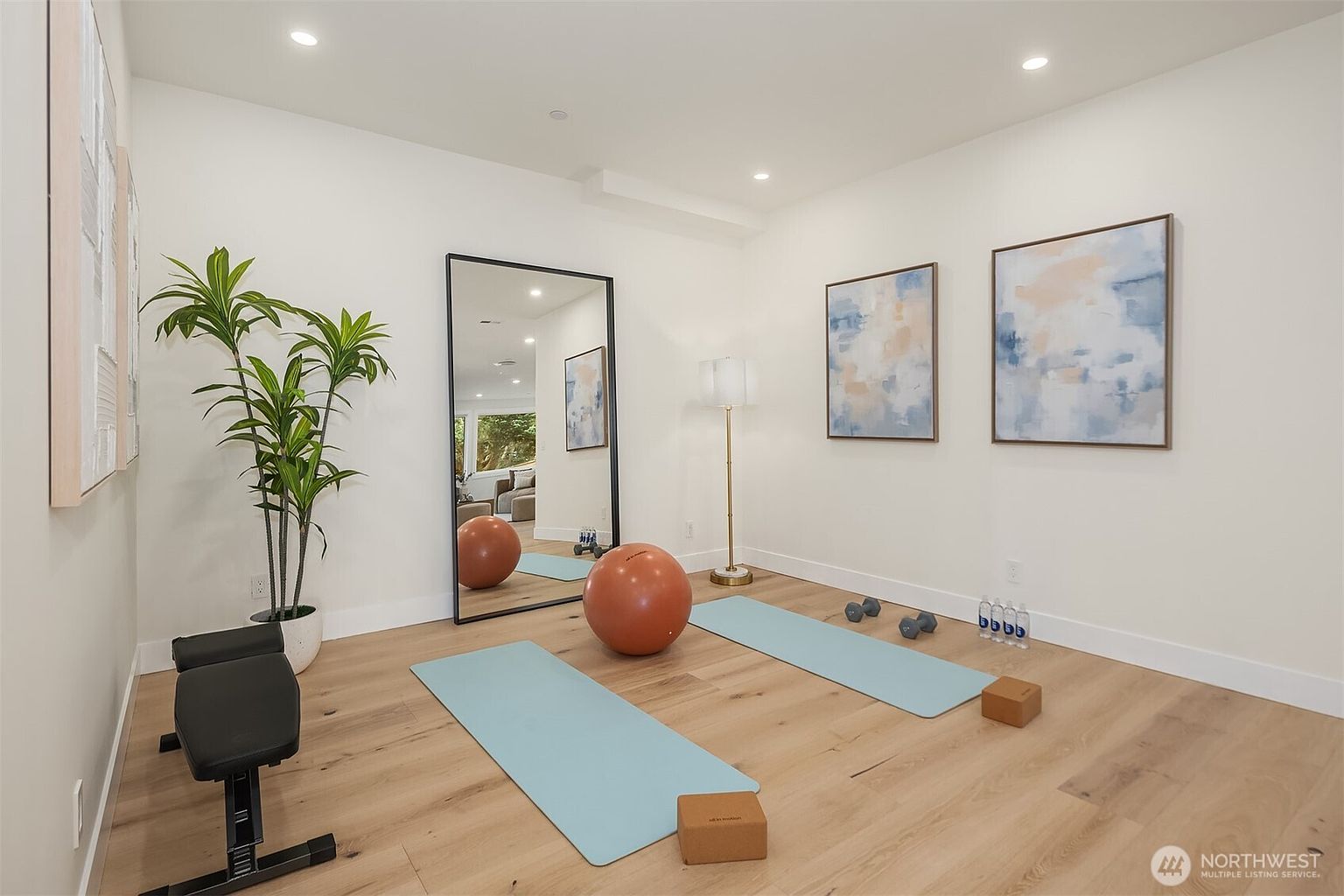 This bright and airy home gym features light wood flooring and crisp white walls, creating a clean, minimalist aesthetic. The space is equipped with two yoga mats, a large exercise ball, dumbbells, yoga blocks, and a weight bench, complemented by a tall floor mirror and two abstract art pieces on the wall. A potted plant adds a touch of greenery, while recessed lighting provides a soft, even glow throughout the room.