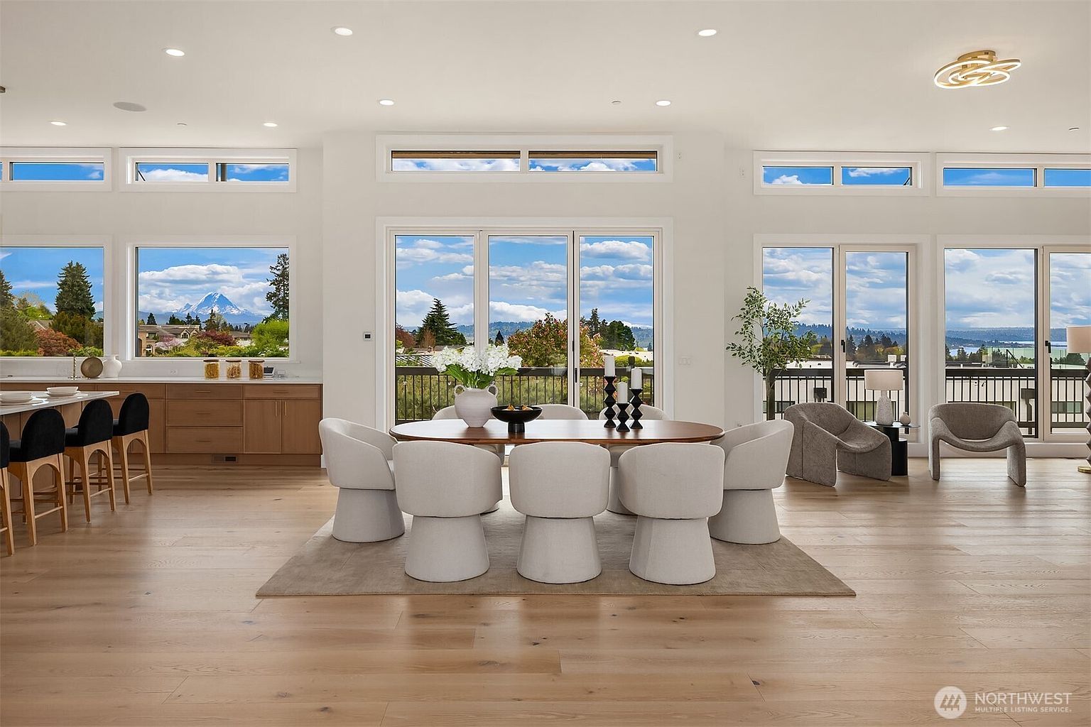 This bright and airy dining area features a large oval table surrounded by six modern, upholstered chairs, set atop a neutral area rug. The space is defined by light wood flooring and expansive windows that provide panoramic views of the landscape and distant mountains. A sleek kitchen island with bar seating is visible to the left, while a pair of contemporary armchairs creates a cozy seating nook on the right, emphasizing an open-concept layout.