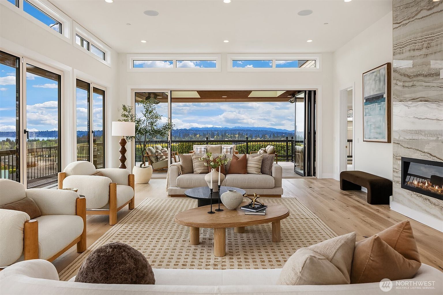 This bright and airy living room features a neutral color palette with plush seating arranged around a central coffee table. Large glass doors open to a spacious deck, seamlessly blending the indoor space with the expansive mountain and water views beyond. A tall stone-clad chimney breast anchors the room, while light wood flooring and recessed lighting contribute to a sophisticated, contemporary aesthetic.