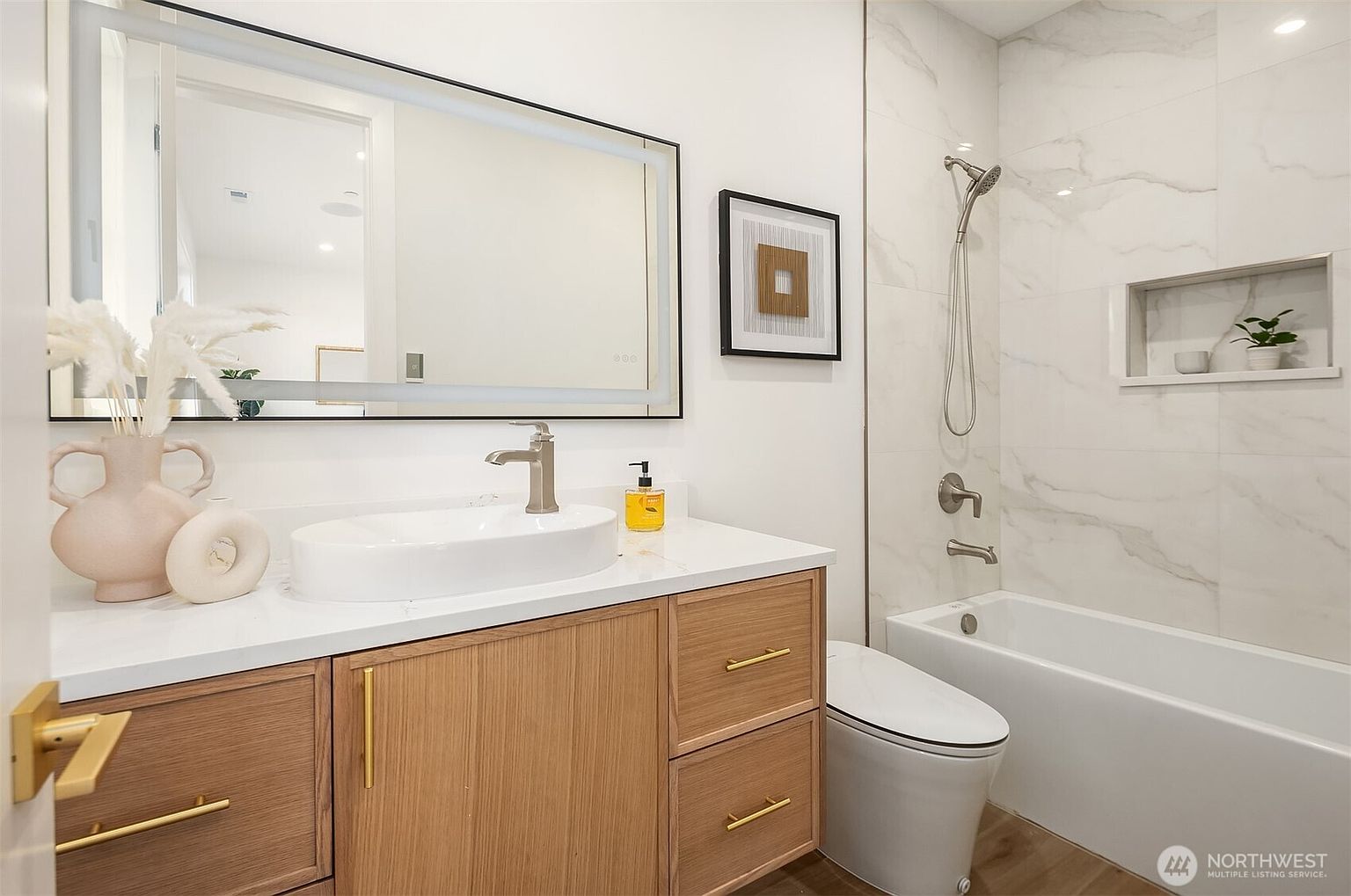 This modern bathroom features a light wood vanity with a vessel sink and a large, rectangular mirror with integrated lighting. To the right, a bathtub is enclosed by white, veined wall tiles, which also feature a convenient recessed niche for storage. The space is bright and clean, accented by gold-toned hardware and a framed piece of wall art.