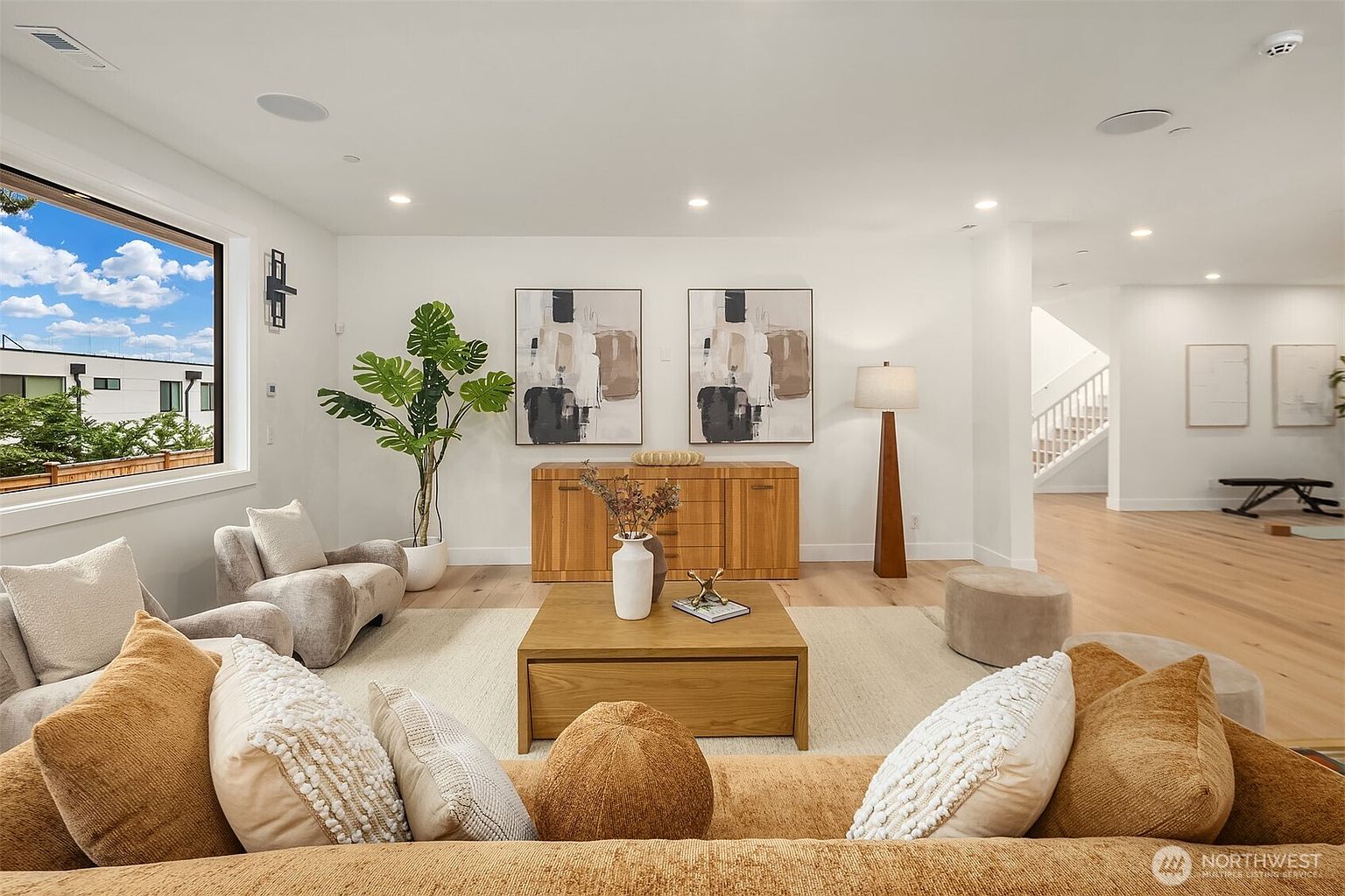 This bright and airy living room features a neutral color palette with warm wood accents and comfortable, textured seating. A large window provides ample natural light, while a wooden console table adorned with abstract art serves as a focal point against the back wall. The space is thoughtfully arranged with a central coffee table and a large area rug, creating an inviting and contemporary atmosphere.