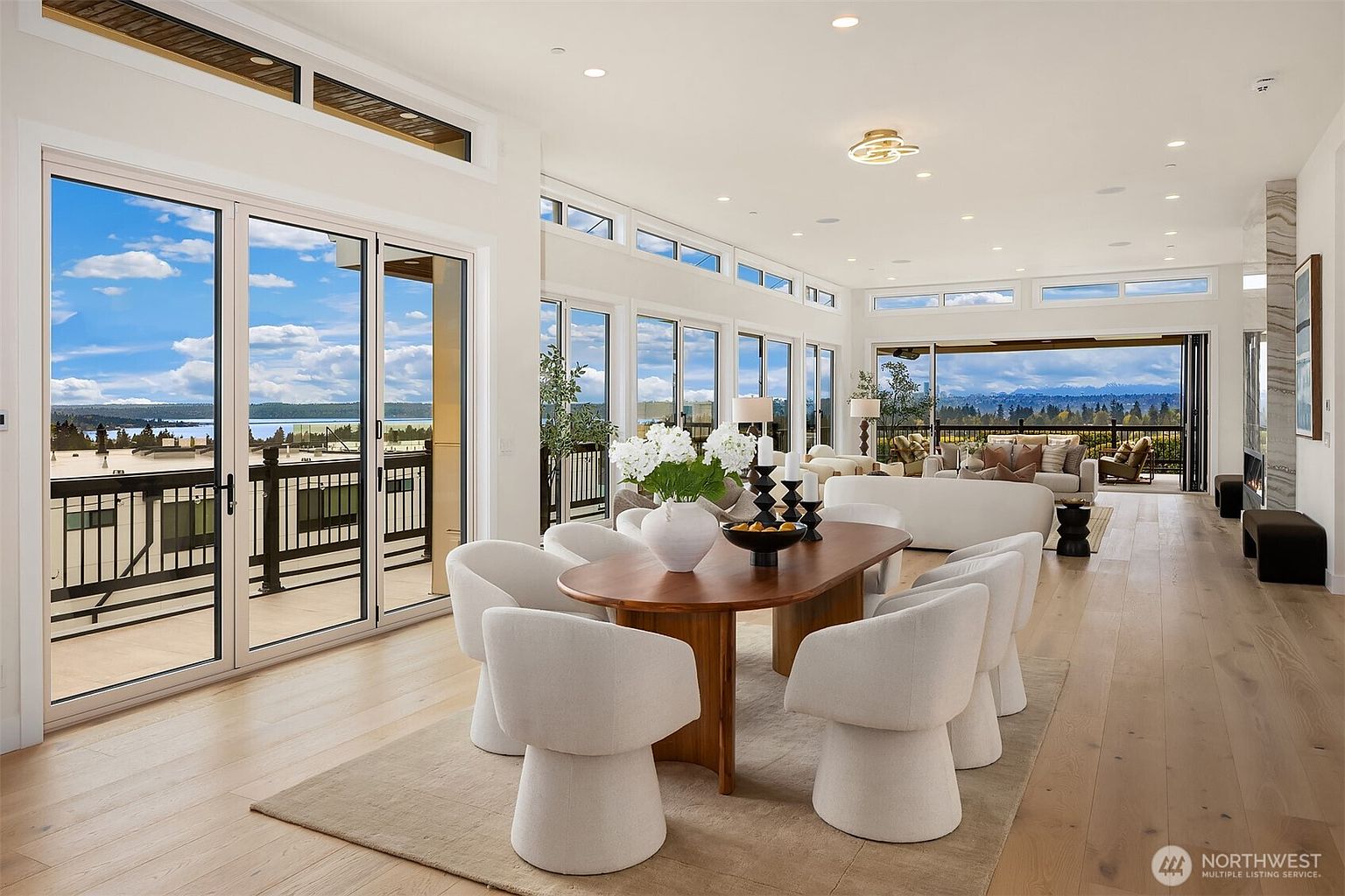 This bright, open-concept dining area features a modern oval wooden table surrounded by six cream-colored upholstered chairs, all set upon a neutral area rug. The space is defined by light wood flooring and an expansive wall of glass doors that provide seamless access to an outdoor deck with scenic water views. Beyond the dining area, the living room continues the airy aesthetic, characterized by minimalist decor and large windows that flood the interior with natural light.