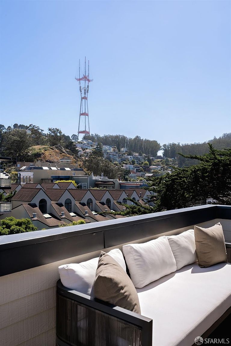 This image captures a private balcony featuring a modern outdoor sofa with plush white cushions and neutral-toned throw pillows. The vantage point offers a sweeping, elevated view of a residential neighborhood with a prominent radio tower rising above the hillside in the distance. The scene conveys a serene and sophisticated atmosphere, perfect for relaxing while taking in the expansive urban landscape.