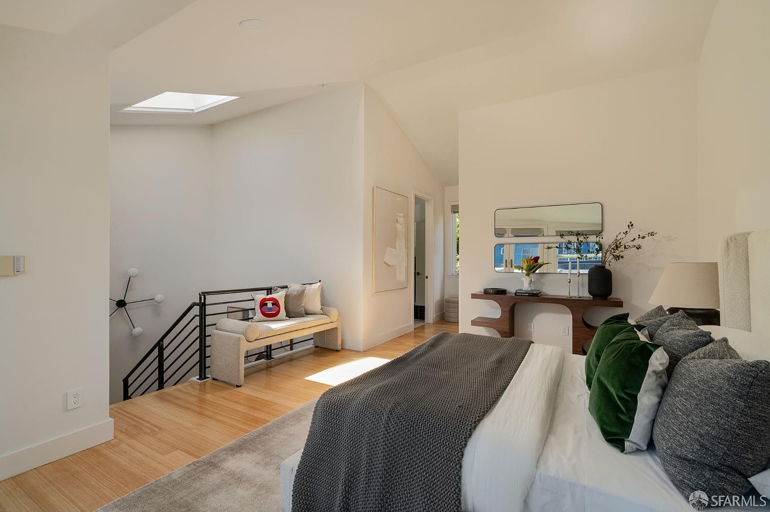 This bright and airy primary bedroom features a vaulted ceiling with a skylight, creating an open and spacious atmosphere. The room includes a cozy seating area near the staircase, a modern console table with dual mirrors, and a plush bed layered with textured linens and green accent pillows. Light wood flooring and neutral walls provide a clean, contemporary aesthetic that feels both sophisticated and inviting.