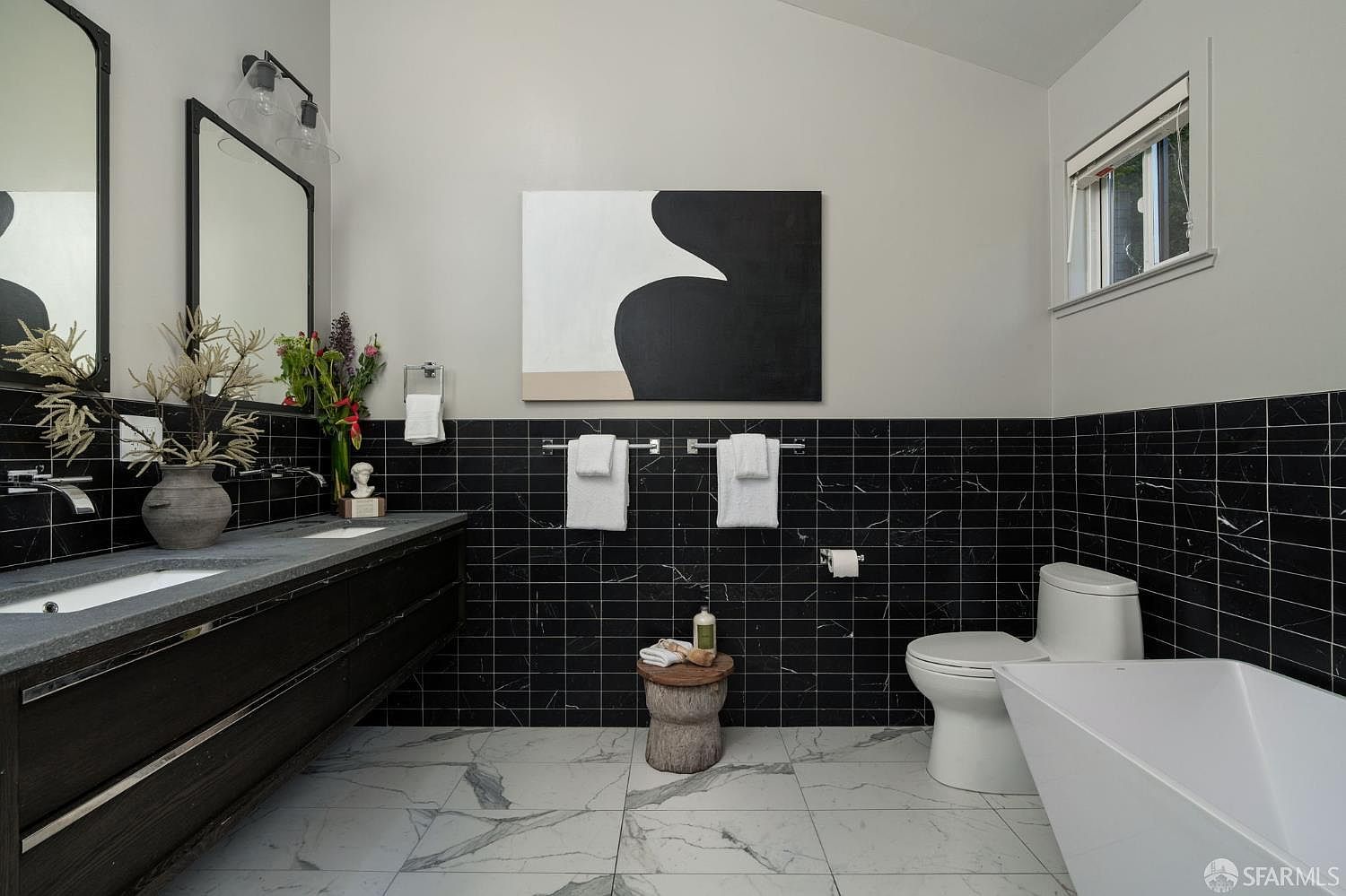 This modern bathroom features a striking contrast between black horizontal subway-style wall tiles and large-format white patterned floor tiles. A long, dark wood vanity with a stone countertop sits beneath a large mirror, while a freestanding white soaking tub is positioned to the right. The space is accented by minimalist fixtures, a wooden stool, and a bold abstract painting, creating a sophisticated and contemporary aesthetic.