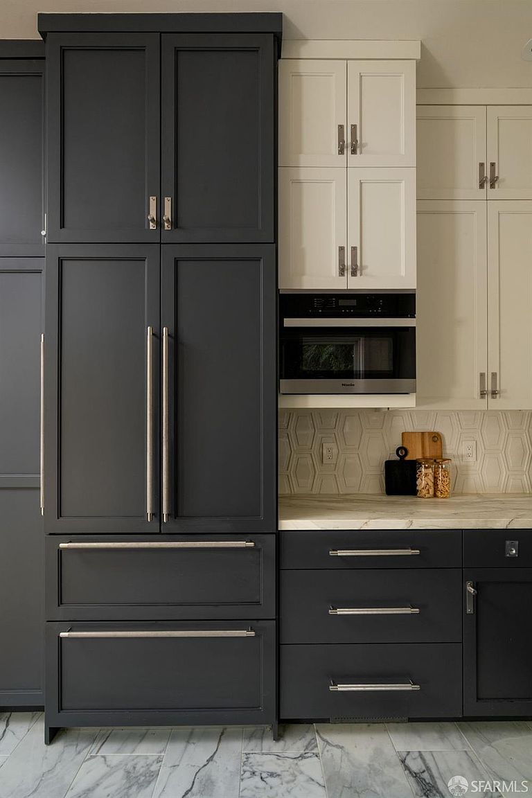 This kitchen features a sophisticated two-tone color palette with dark charcoal cabinetry paired with contrasting white upper cabinets. The space includes a built-in refrigerator, a wall-mounted microwave, and a patterned tile backsplash above a stone countertop. The perspective is a straight-on, eye-level shot that highlights the clean lines and modern hardware of the cabinetry.