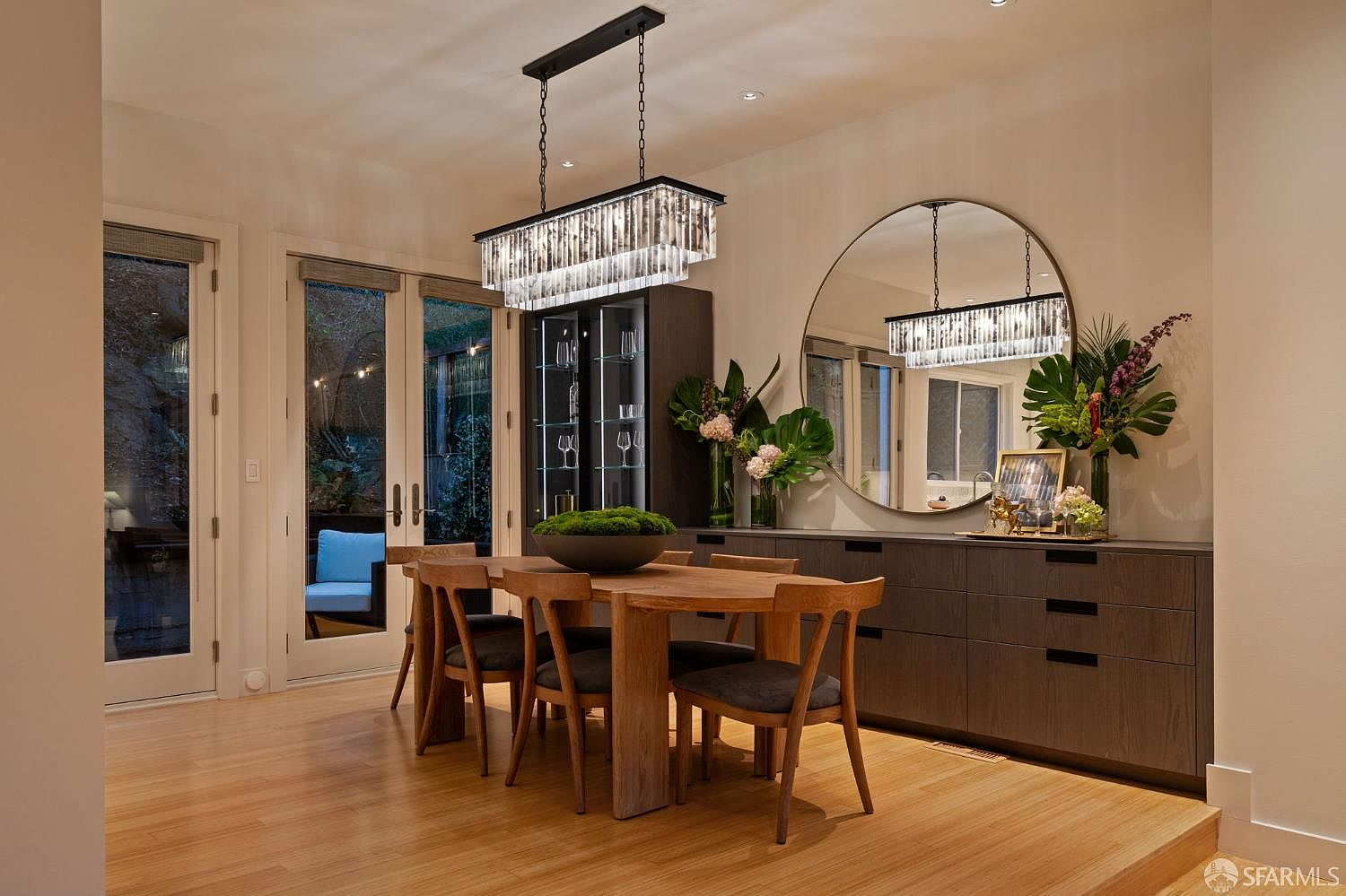 This elegant dining room features a warm wood dining table surrounded by chairs with dark upholstered seats, centered beneath a sophisticated rectangular crystal chandelier. A large, circular mirror hangs above a long, dark wood sideboard, reflecting the dining area and adding depth to the space. To the left, glass-paned doors provide a view of the outdoor area, while a built-in glass-front display cabinet adds a refined touch to the room's contemporary aesthetic.