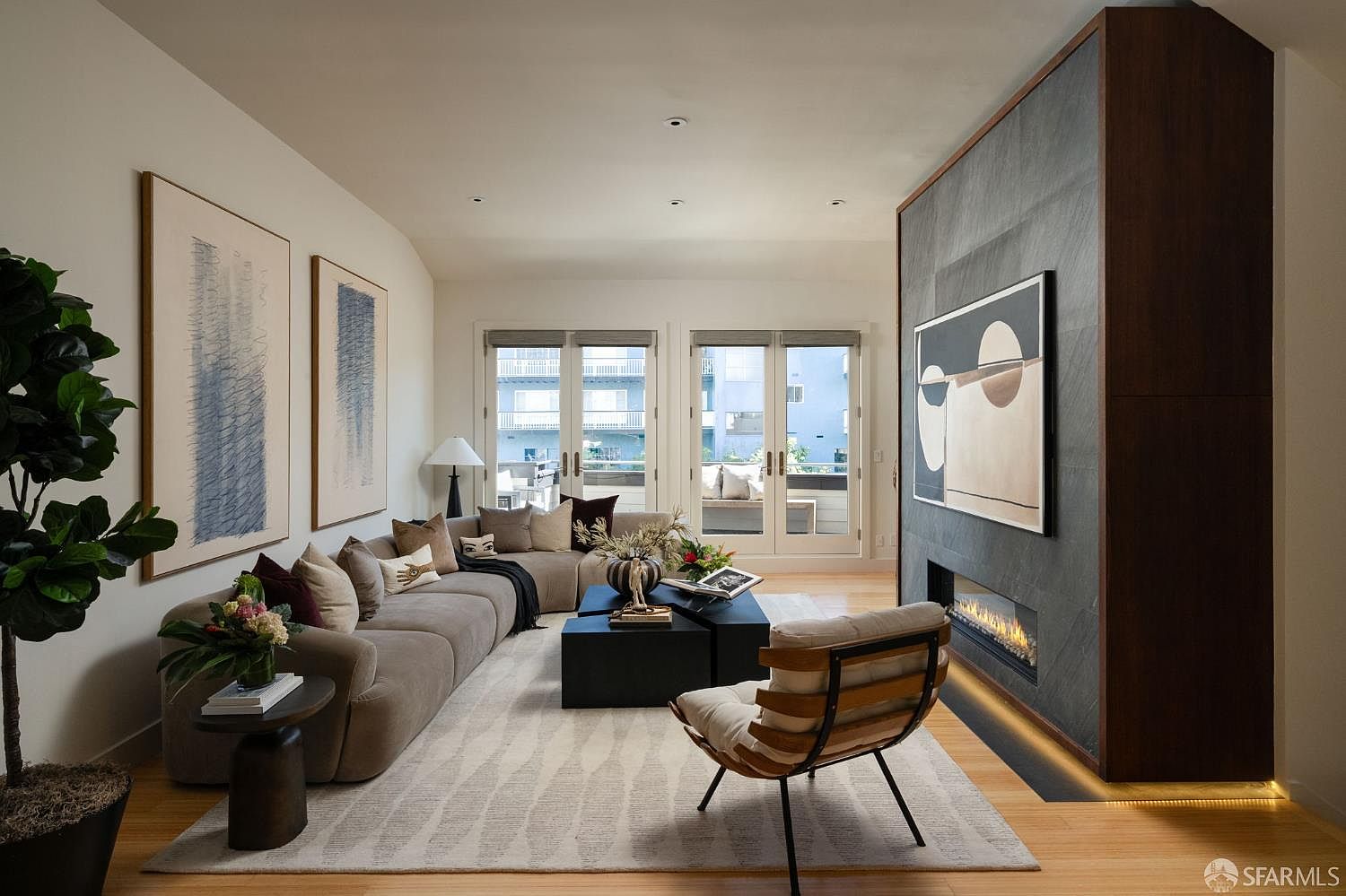 This sophisticated living room features a large, plush sectional sofa arranged around a set of dark, geometric coffee tables on a textured area rug. A striking stone-clad chimney breast serves as a focal point, housing a linear fireplace and a piece of modern abstract art. The space is brightened by natural light streaming through two sets of glass doors that lead to an outdoor balcony, creating a seamless transition between indoor and outdoor living.