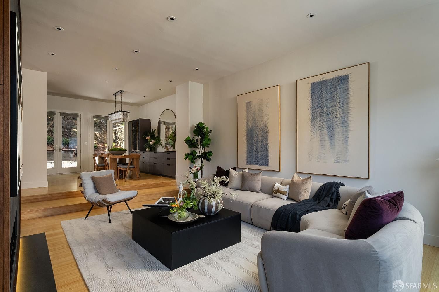 This sophisticated living room features a large, plush sectional sofa arranged around a minimalist black coffee table, set atop a textured area rug. The space is accented by two large abstract art pieces on the wall and a modern lounge chair, with a raised dining area visible in the background through an open-concept layout. The room is bathed in natural light from the rear glass doors, creating an airy and inviting atmosphere with a contemporary aesthetic.
