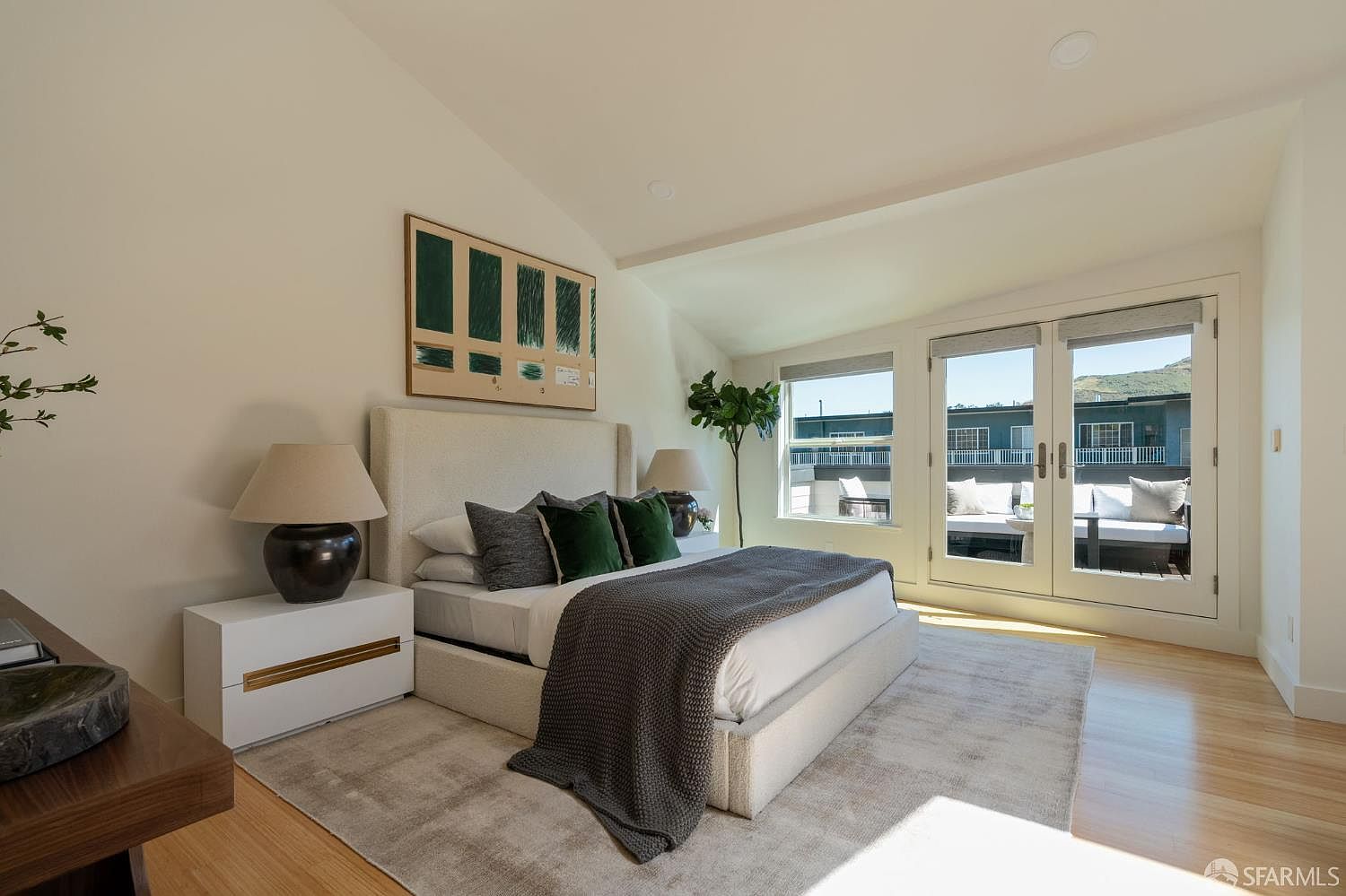 This primary bedroom features a neutral color palette with a large upholstered bed as the focal point, accented by dark green throw pillows and a textured gray throw blanket. The room is illuminated by two bedside lamps on modern white nightstands and benefits from natural light streaming through large glass doors that open onto a private balcony. The space is finished with light wood flooring and a large area rug, creating a serene and contemporary atmosphere.