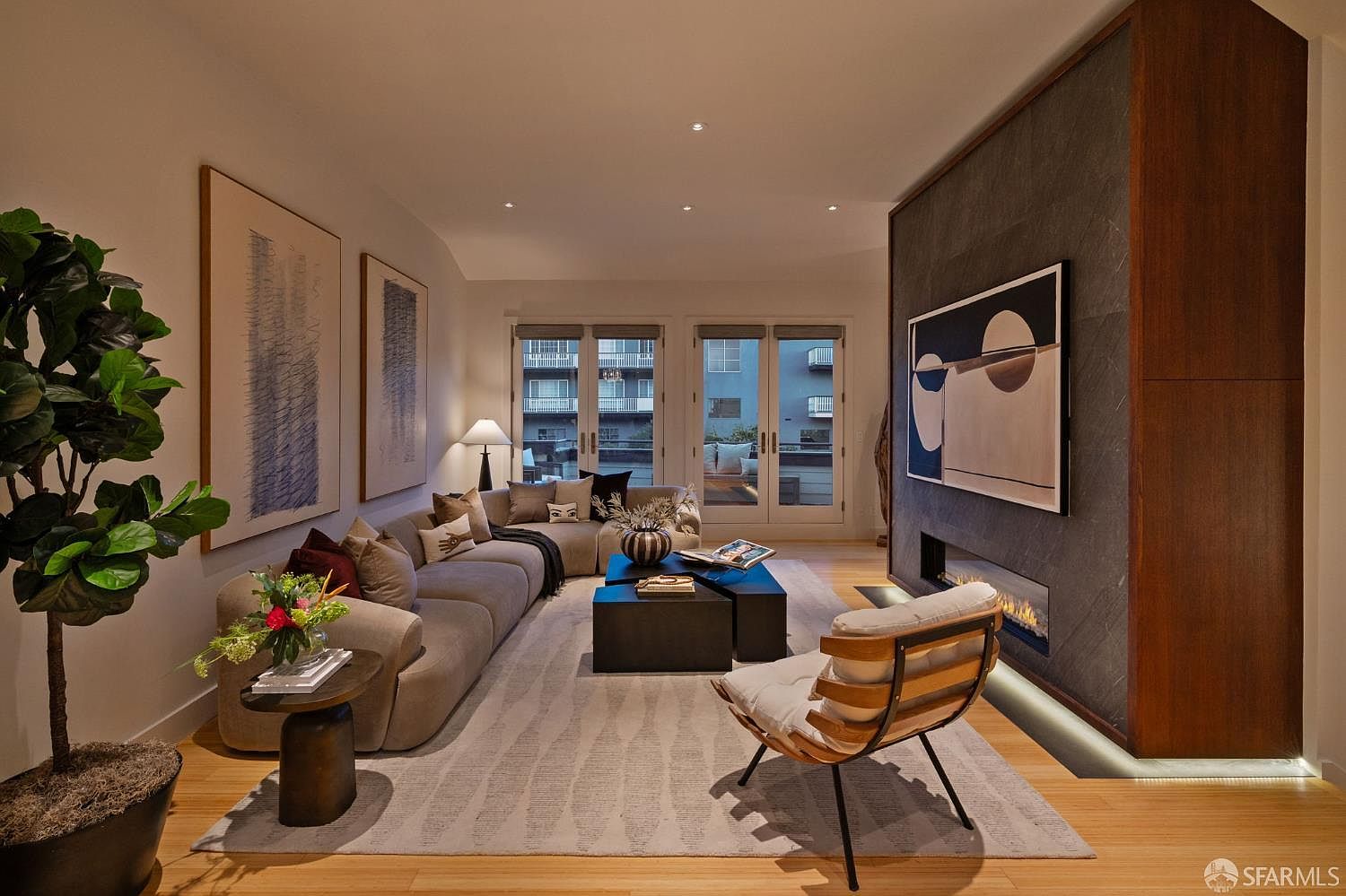 This sophisticated living room features a large, plush sectional sofa arranged around a set of dark, modern coffee tables on a textured area rug. A striking focal point is the dark stone-clad chimney breast housing a linear fireplace, complemented by a large abstract art piece above. The space is illuminated by recessed lighting and natural light streaming through glass doors that lead to an outdoor balcony, creating an inviting and contemporary atmosphere.