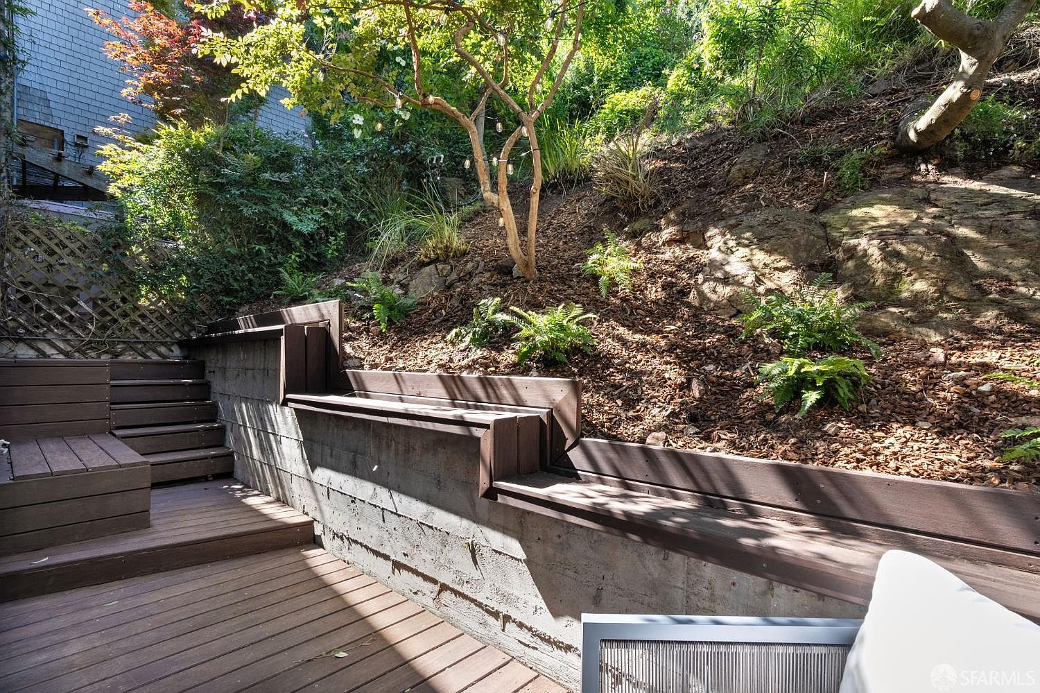 This outdoor space features a multi-level wooden deck integrated with a concrete retaining wall that doubles as built-in bench seating. The area transitions into a sloped, natural hillside landscape filled with mulch, ferns, and a mature tree adorned with string lights. The perspective is from a low angle, emphasizing the tiered structure and the blend of modern decking with the rugged, natural terrain.