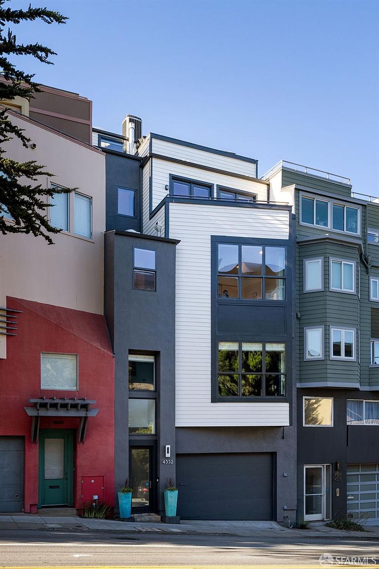 This contemporary multi-story townhouse features a striking facade with a mix of dark gray stucco, white horizontal siding, and a bold red accent wall on the adjacent unit. The home includes a private garage, a modern glass-paneled front door, and large windows that provide a clean, urban aesthetic. The perspective is a straight-on street-level shot, capturing the architectural contrast between the modern design and the neighboring structures.