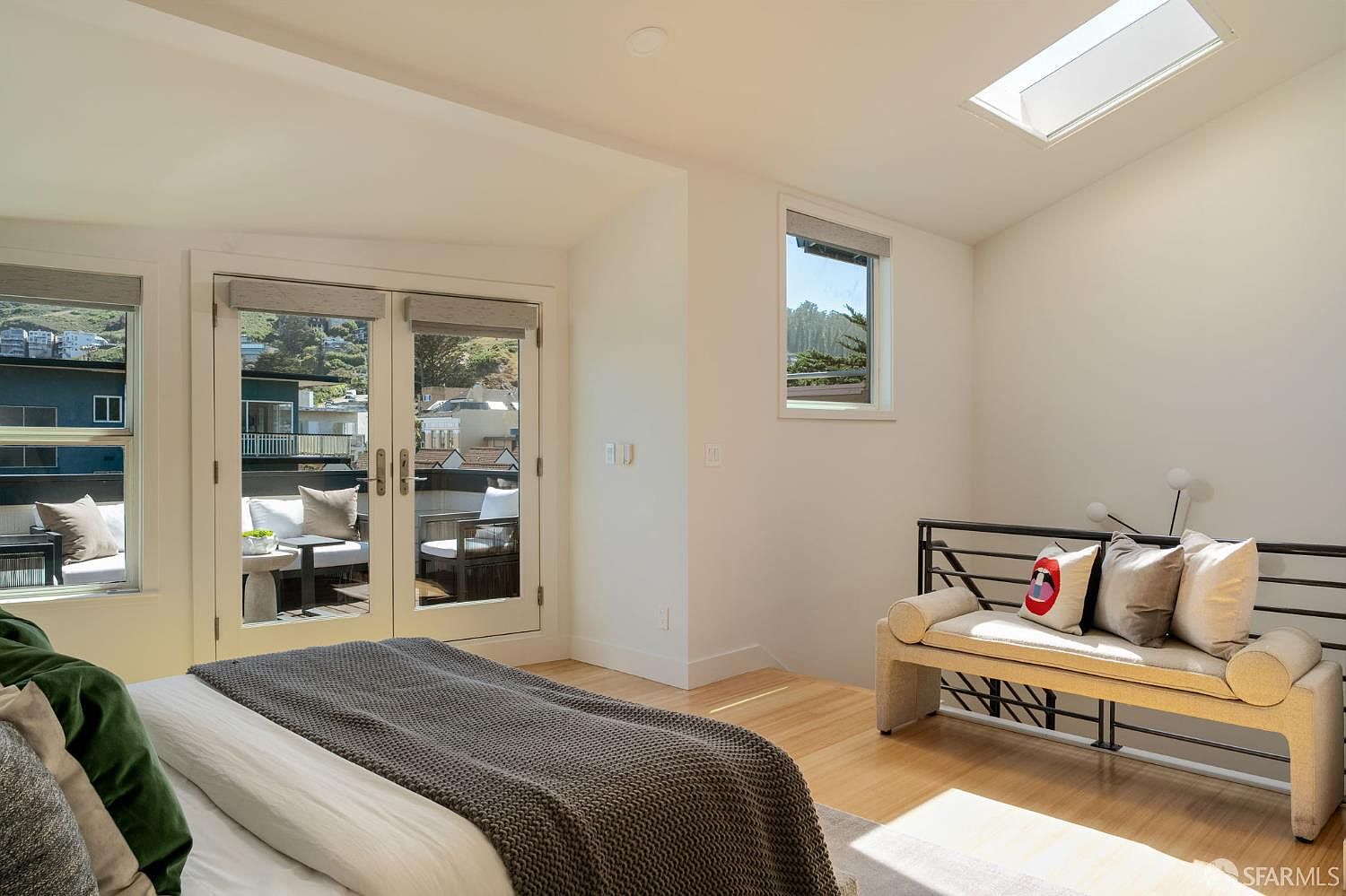 This bright and airy primary bedroom features light wood flooring and a vaulted ceiling with a skylight that floods the space with natural light. A set of French doors opens onto a private balcony, while a stylish bench with decorative pillows sits against a metal-framed railing. The room offers a clean, modern aesthetic with neutral walls and a comfortable, inviting atmosphere.