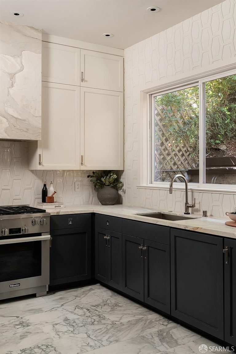 This modern kitchen features a high-contrast design with dark lower cabinetry paired with crisp white upper cabinets and a stone-clad range hood. The space is accented by a geometric patterned backsplash and light-toned stone countertops, while a window above the sink provides a view of the outdoor greenery. The perspective is a medium-angle shot capturing the functional workspace, highlighting the sleek stainless steel appliances and marble-patterned flooring.