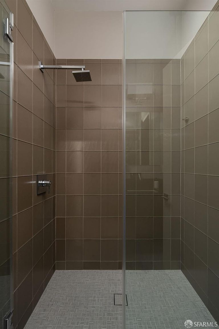 This modern walk-in shower features large, neutral-toned square tiles on the walls and a textured, small-format patterned tile on the floor. A sleek, chrome rainfall showerhead is mounted on the wall, complemented by a minimalist control handle. The space is enclosed by a clear glass panel, creating an open and airy feel within the shower stall.
