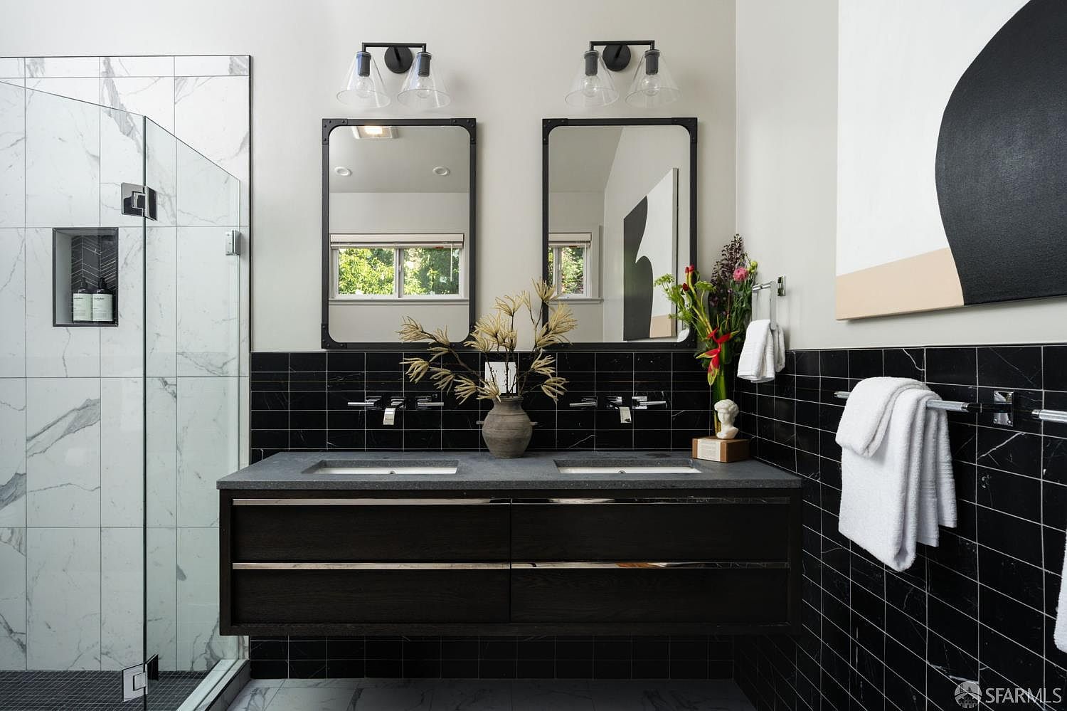This modern primary bathroom features a floating double vanity with a dark wood finish and a stone countertop, complemented by dual rectangular mirrors and industrial-style wall sconces. The walls are adorned with black horizontal tiles, creating a striking contrast against the white marble-patterned shower enclosure to the left. A decorative vase with dried botanicals and a small bust sculpture add a sophisticated, curated touch to the vanity space.
