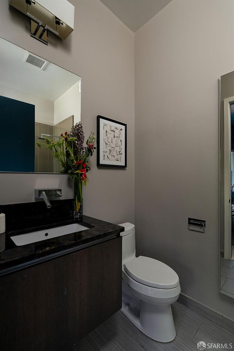 This modern powder room features a sleek, dark-toned vanity with a stone countertop and a wall-mounted faucet. A large, frameless mirror reflects the space, which is accented by a framed piece of abstract art and a vibrant floral arrangement. The room is finished with neutral gray walls and a white toilet, creating a clean and sophisticated aesthetic.