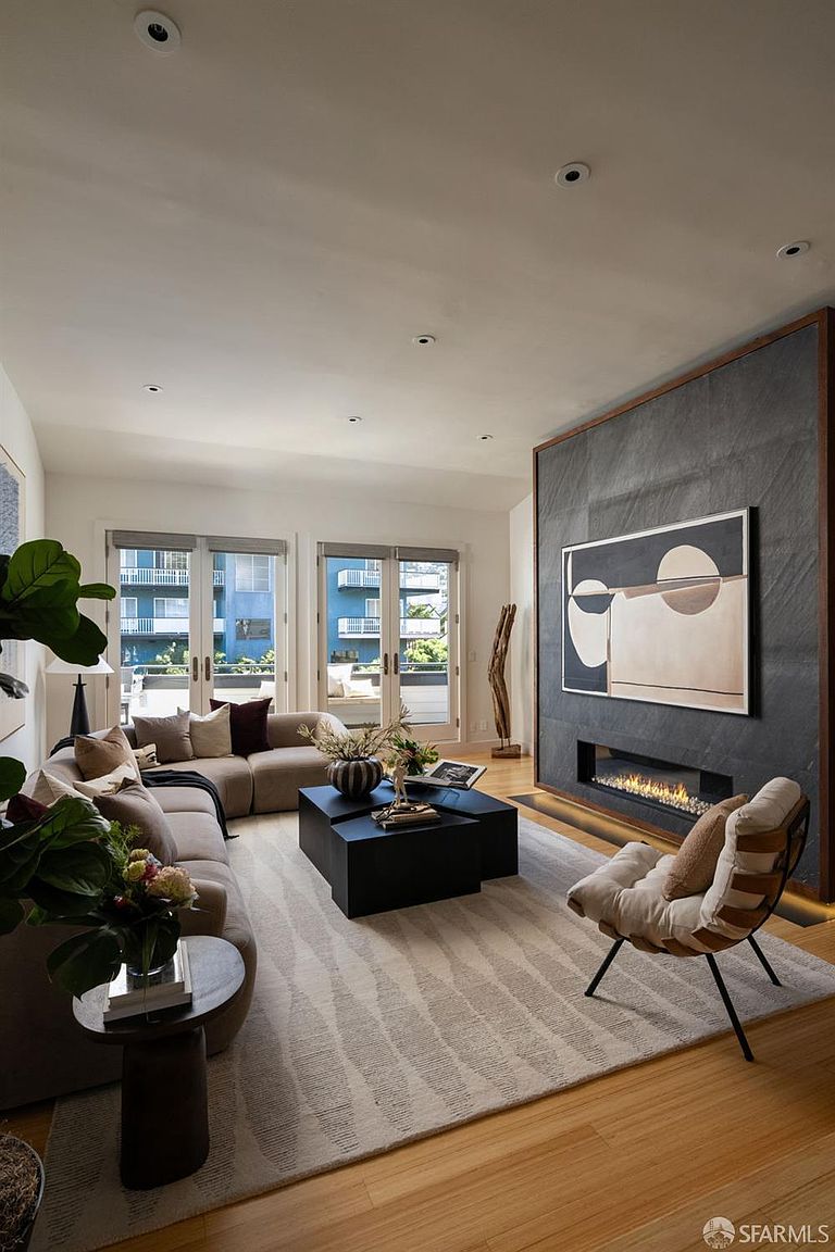 This sophisticated living room features a striking dark stone-clad chimney breast that serves as a focal point, housing a linear fireplace and a large piece of abstract art. The space is furnished with a plush, neutral-toned sectional sofa, a set of modular black coffee tables, and a modern accent chair, all arranged on a textured area rug over light wood flooring. Large glass doors provide ample natural light and a view of the exterior balcony, creating an airy and inviting atmosphere.
