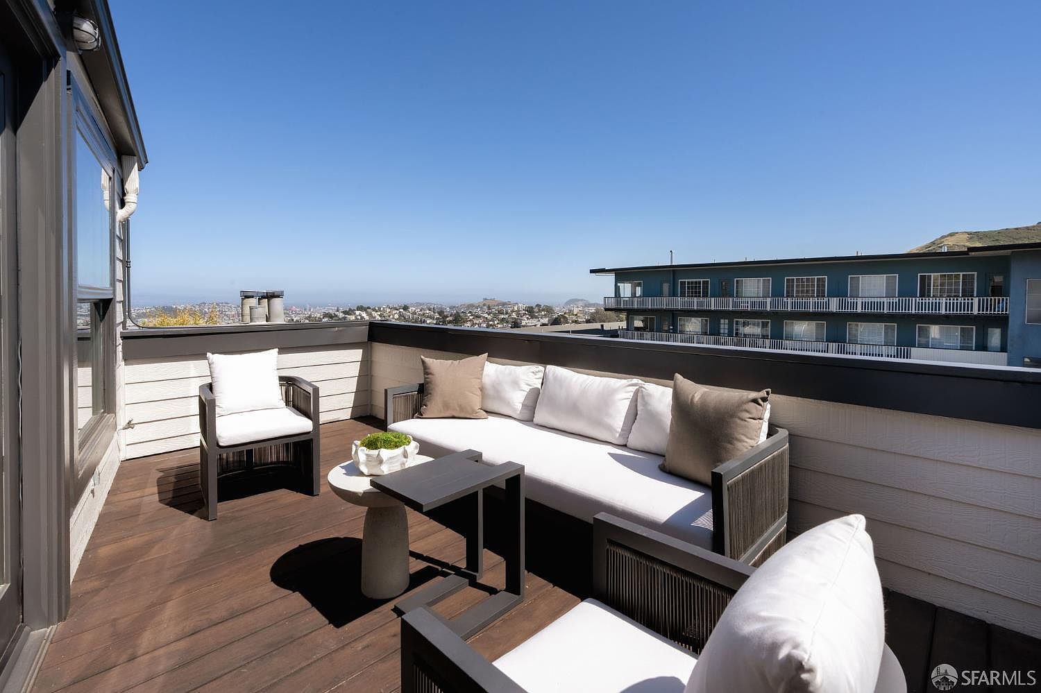 This elevated outdoor deck features a modern seating arrangement with a long sofa and two armchairs arranged around a small stone-topped table. The space is finished with dark wood decking and offers an expansive, unobstructed view of the surrounding cityscape and distant hills under a clear blue sky. The perspective is from a slightly elevated vantage point, capturing the inviting atmosphere of this private urban retreat.