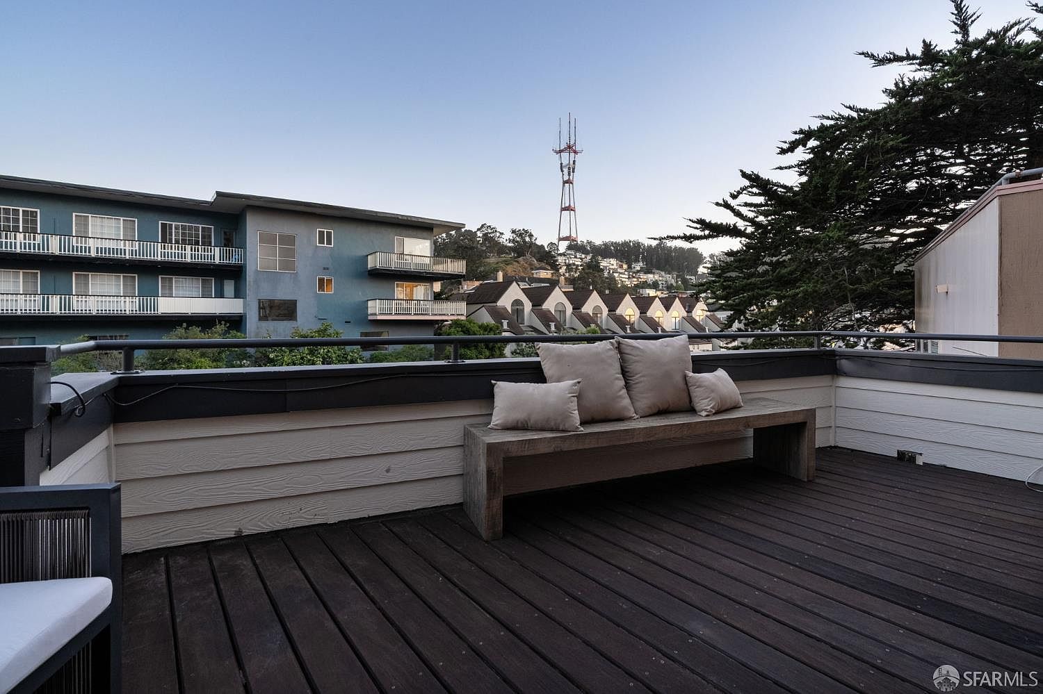 This elevated outdoor deck features dark wood-toned decking and a simple wooden bench adorned with several neutral-toned throw pillows. The space is enclosed by a black metal railing, offering an unobstructed view of the surrounding neighborhood and the iconic Sutro Tower in the distance. The perspective is from a slightly elevated vantage point, capturing the serene atmosphere of this private urban retreat.