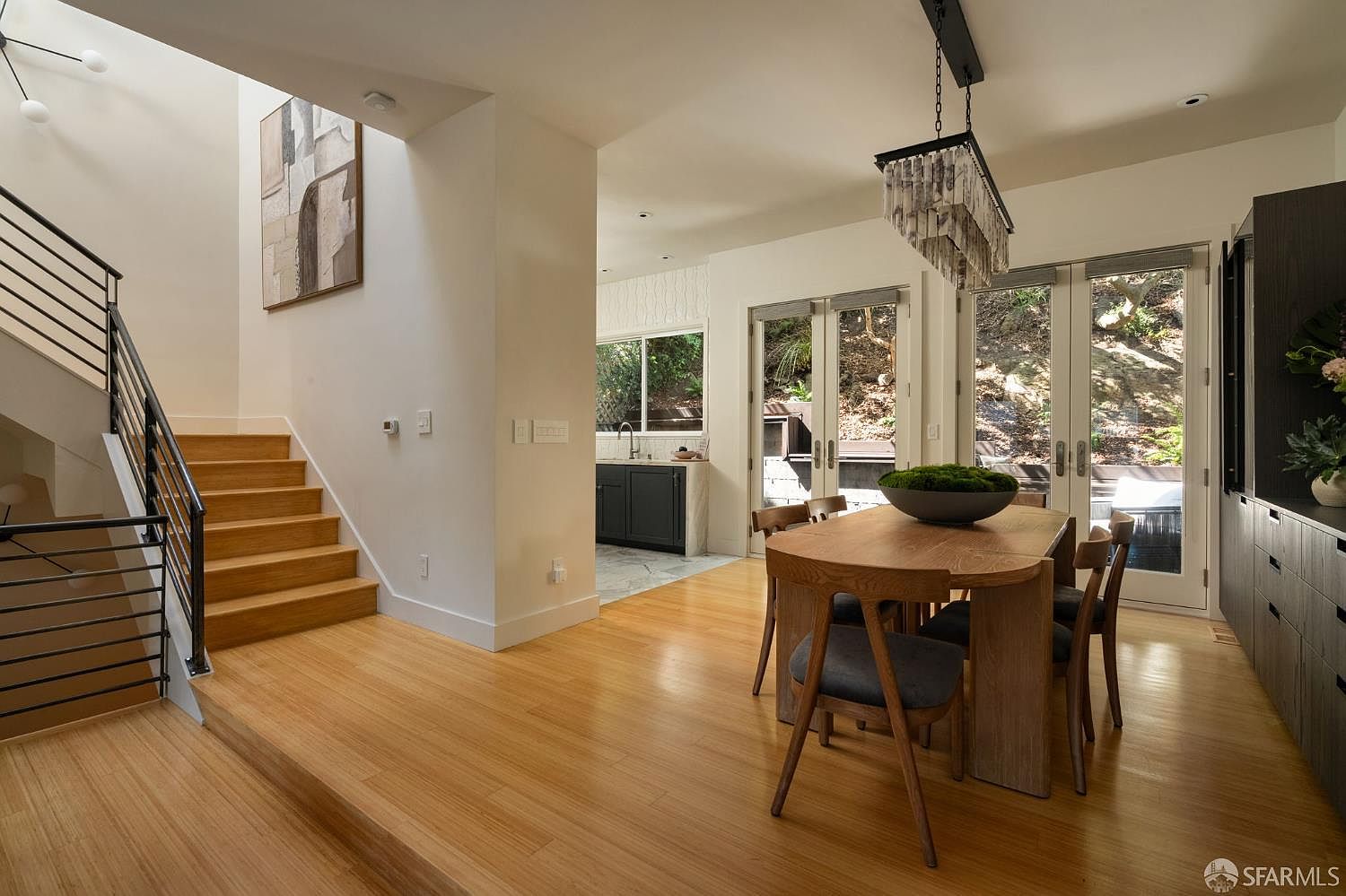This bright and open dining area features light wood flooring and a modern wooden dining table centered under a unique, tiered chandelier. To the left, a staircase with a black metal railing leads upward, while a doorway in the background reveals a glimpse of a kitchen with dark cabinetry. Large glass doors provide a view of the wooded exterior, filling the space with natural light and creating a seamless transition between indoor and outdoor living.