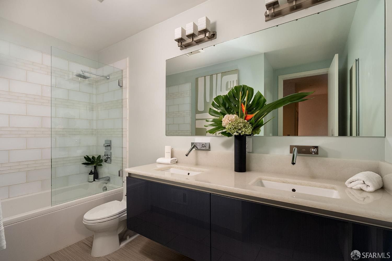 This modern primary bathroom features a sleek, dark-toned floating double vanity topped with a light-colored stone counter and wall-mounted faucets. A large, frameless mirror spans the width of the vanity, reflecting a glass-enclosed shower area with light-colored subway tile walls. The space is accented by a tall, contemporary light fixture above the mirror and a vibrant floral arrangement, creating a clean and sophisticated aesthetic.