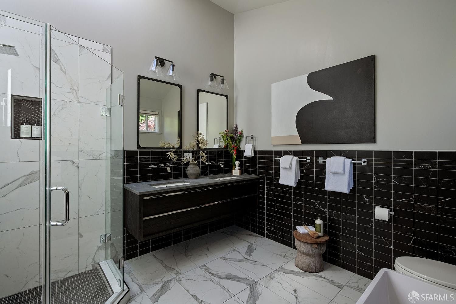 This modern primary bathroom features a striking contrast between black subway-style wall tiles and large-format white patterned floor tiles. The space includes a double vanity with a dark wood cabinet, a glass-enclosed shower with a niche, and a minimalist aesthetic accented by a large abstract art piece. The perspective captures the vanity area and shower, highlighting the clean lines and contemporary design choices.