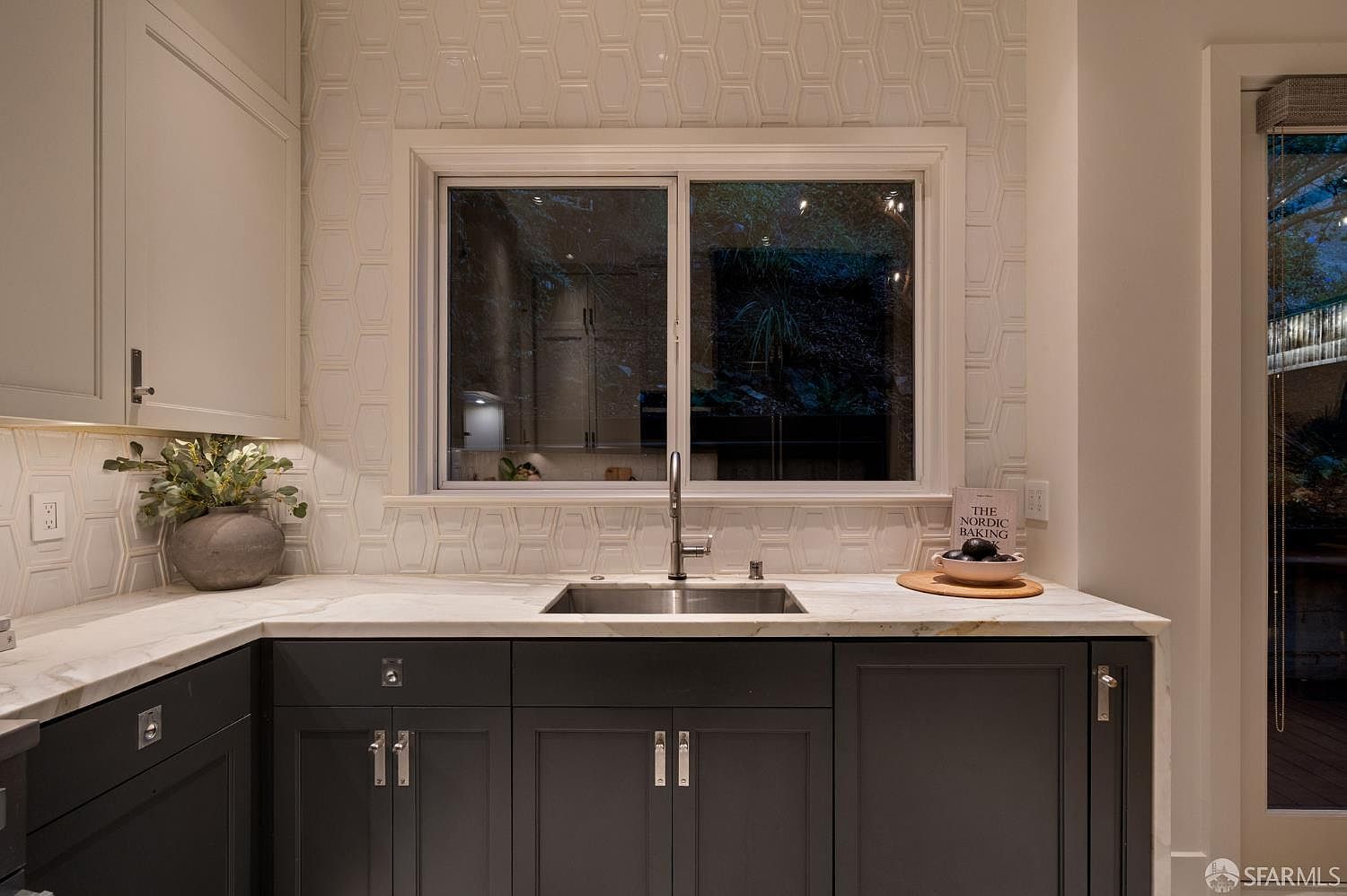 This kitchen area features dark charcoal cabinetry paired with a light-toned stone countertop and a unique geometric patterned tile backsplash. A central window provides a view to the exterior, while a decorative bowl and a cookbook add a touch of personality to the workspace. The space is illuminated by under-cabinet lighting, creating a sophisticated and functional atmosphere.