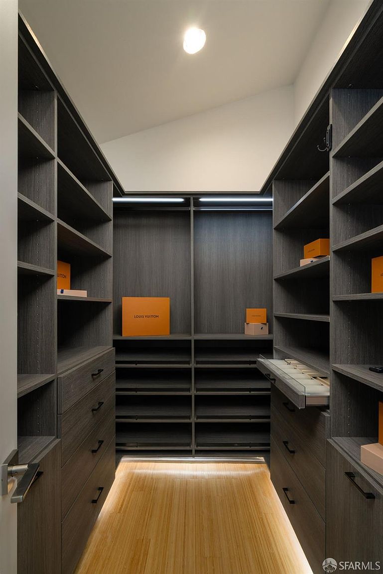 This walk-in closet features custom dark wood-toned cabinetry with a mix of open shelving, hanging rods, and drawers. The space is illuminated by integrated LED lighting strips and a single ceiling light, highlighting the light-toned wood flooring. The symmetrical layout provides a clean, organized, and luxurious aesthetic suitable for high-end storage.