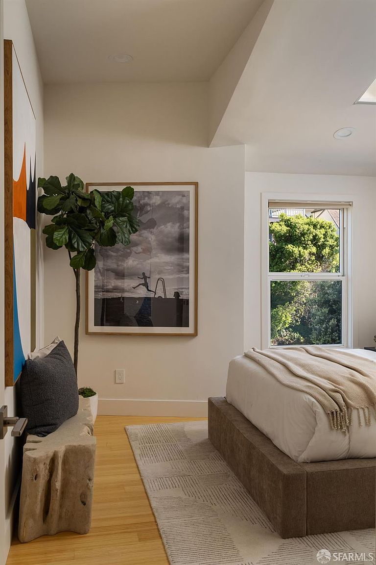 This serene bedroom features a neutral color palette with a plush, upholstered bed frame and a textured area rug. A large, framed black-and-white photograph hangs on the wall, complemented by a tall potted plant and a rustic, carved wood bench in the foreground. The room is bathed in natural light from a window that offers a view of lush greenery outside, creating a calm and inviting atmosphere.