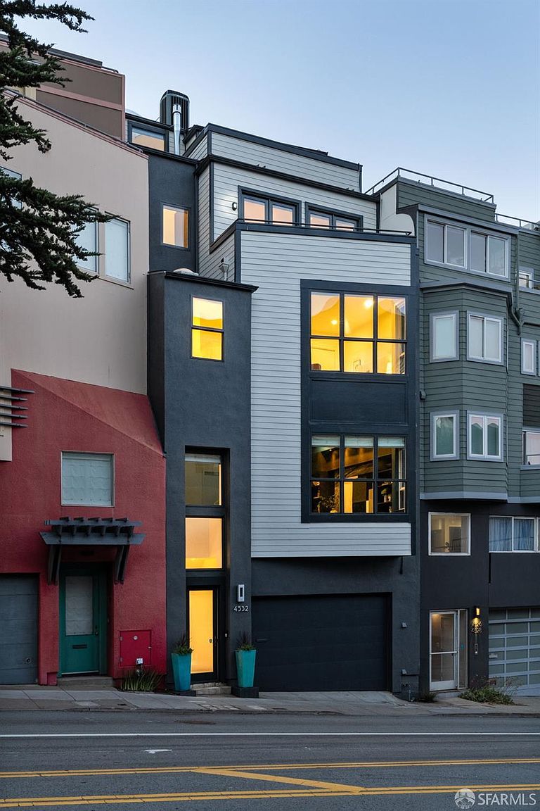 This contemporary multi-level townhouse features a striking facade with a mix of dark gray stucco and light-colored horizontal siding. The structure includes a prominent garage, a vertical entryway with a glass door, and large windows that allow warm light to glow from within. The perspective is a straight-on street view, showcasing the modern architectural design and its integration into the urban streetscape.