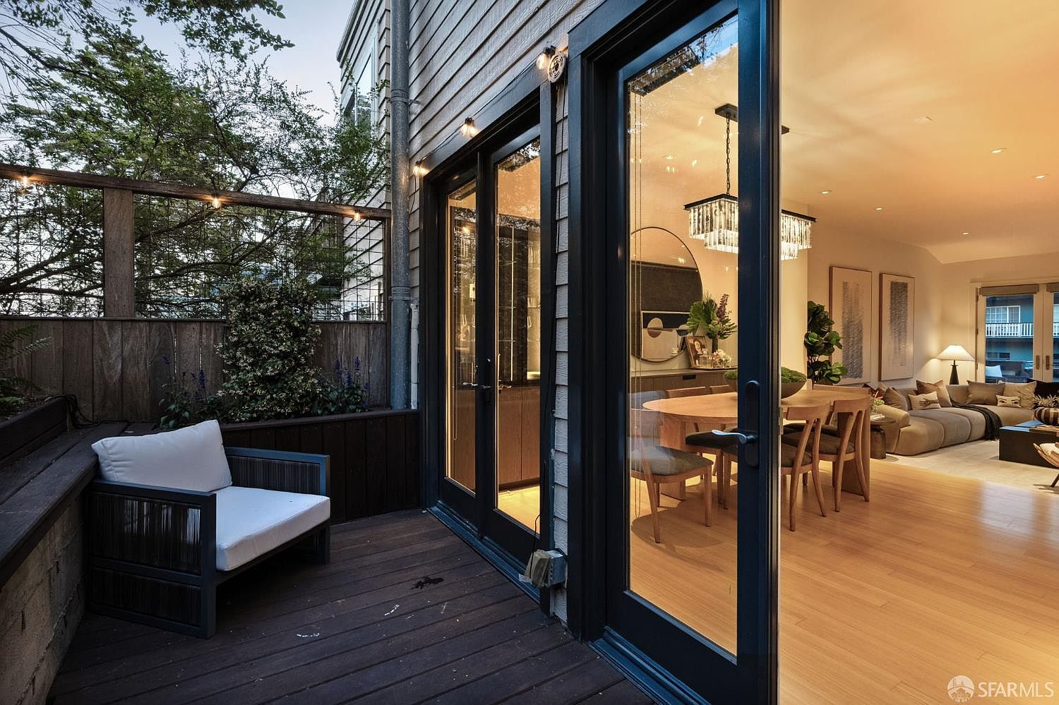 This image captures a seamless transition between a private wooden deck and a bright, open-concept interior living space. The deck features a modern lounge chair and string lighting, while the interior reveals a dining area and a comfortable living room with light wood flooring. Large glass doors provide an expansive view, creating an inviting indoor-outdoor flow perfect for entertaining.
