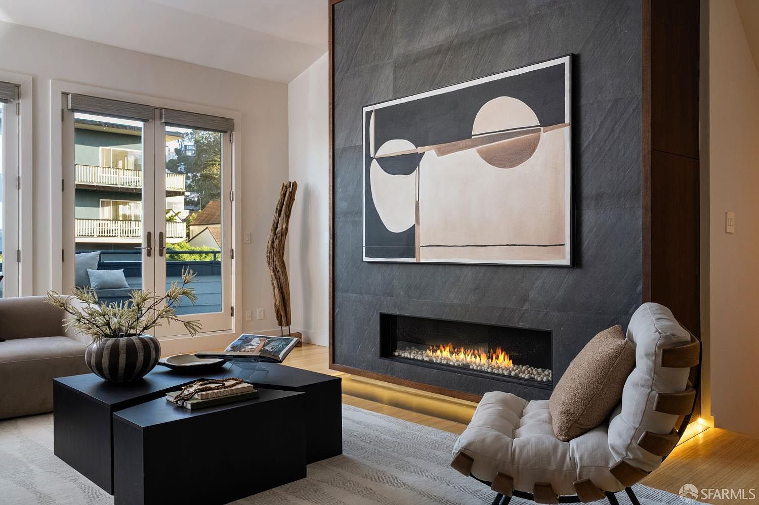This sophisticated living room features a striking dark stone-clad chimney breast that serves as a focal point, housing a sleek linear fireplace. A large abstract painting hangs above the fireplace, while a modern, low-profile lounge chair and minimalist black coffee tables define the seating area. The space is brightened by tall glass doors that open to a balcony, creating a seamless transition between the refined interior and the outdoor view.